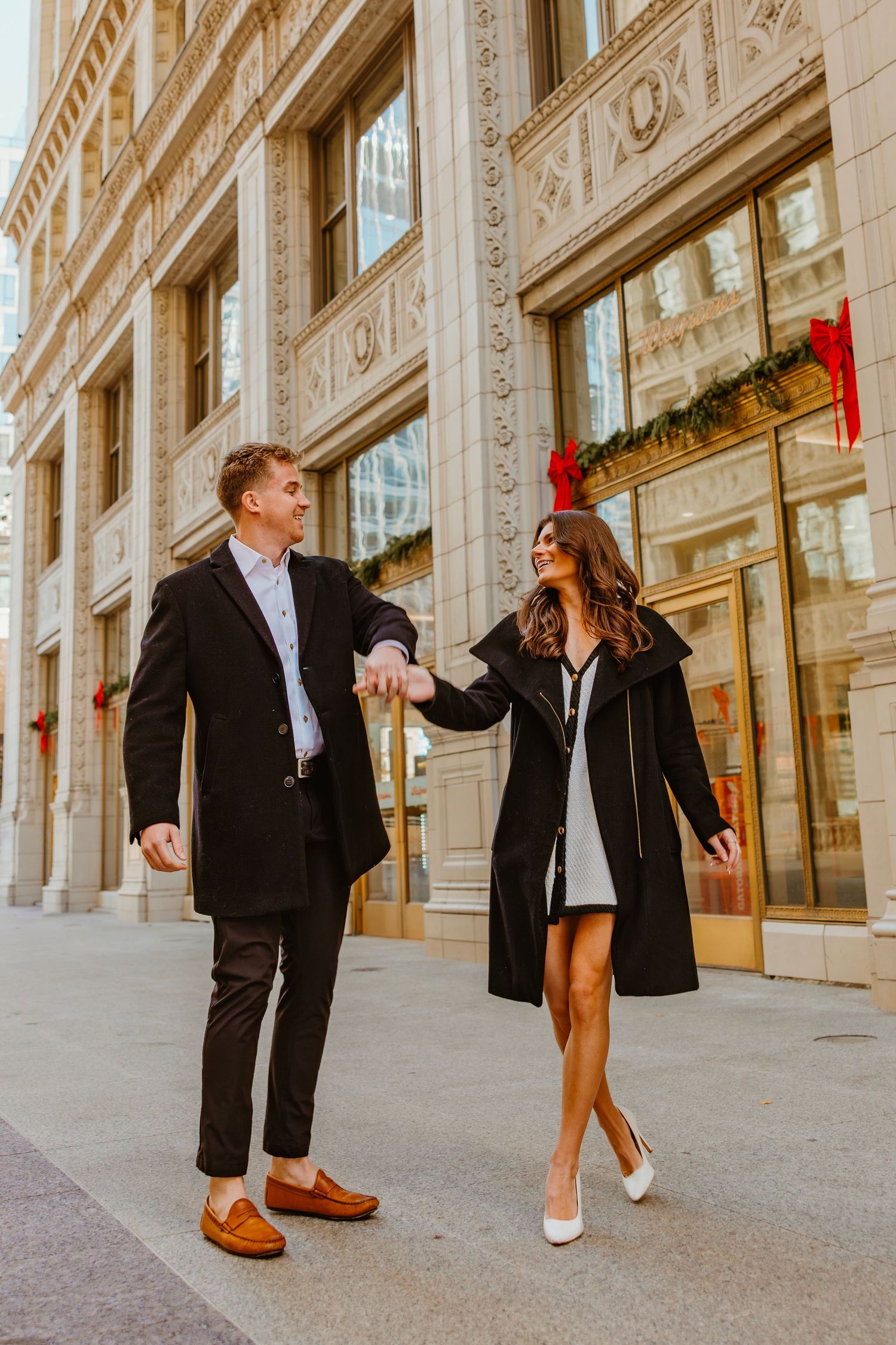 Stylish couple holding hands and walking past a grand building adorned with Christmas decorations. Outdoor Chicago engagement photos.