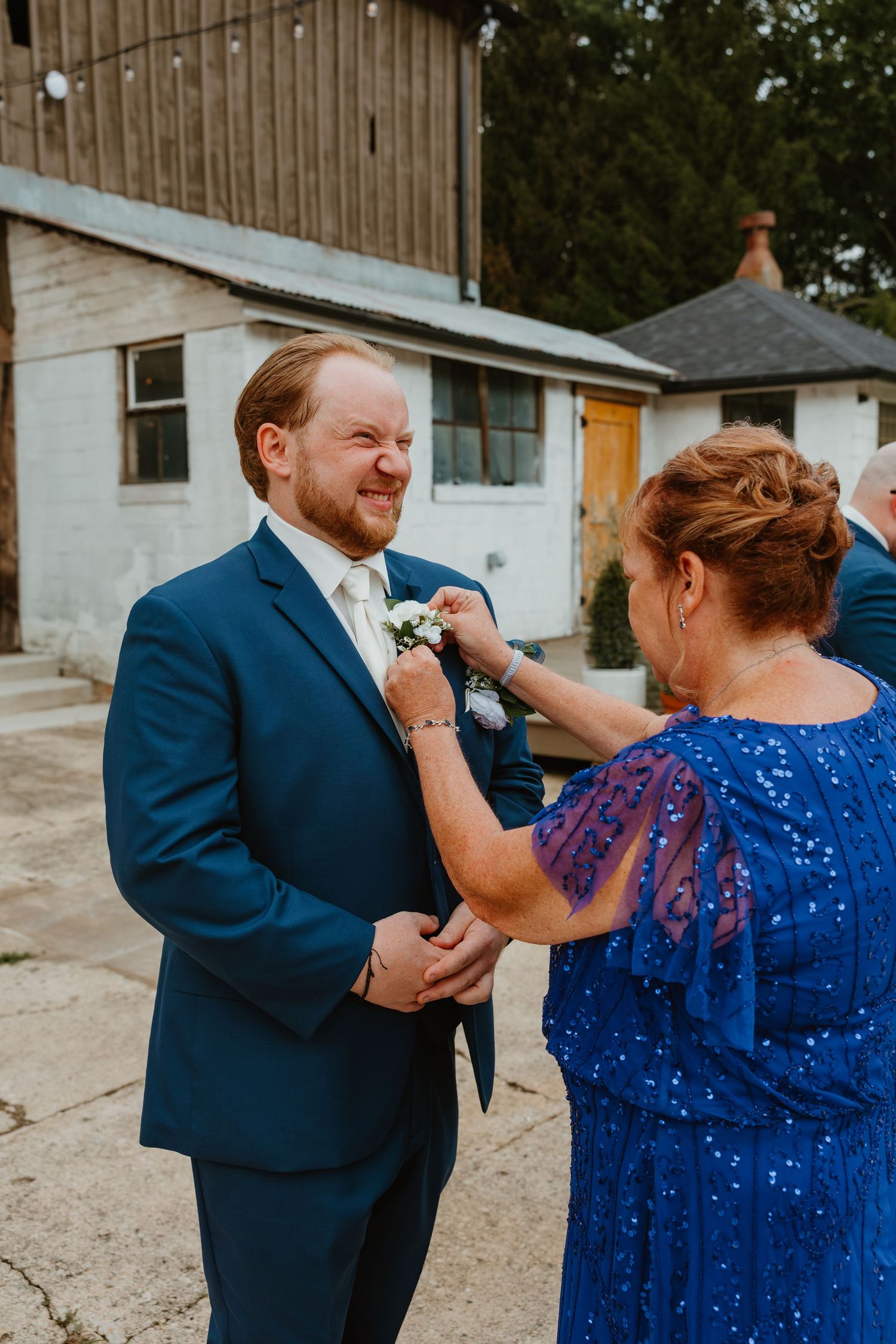 Mother pins a white boutonniere on her smiling groom son in a navy suit outside a rustic barn wedding venue. Northwest Indiana wedding venue