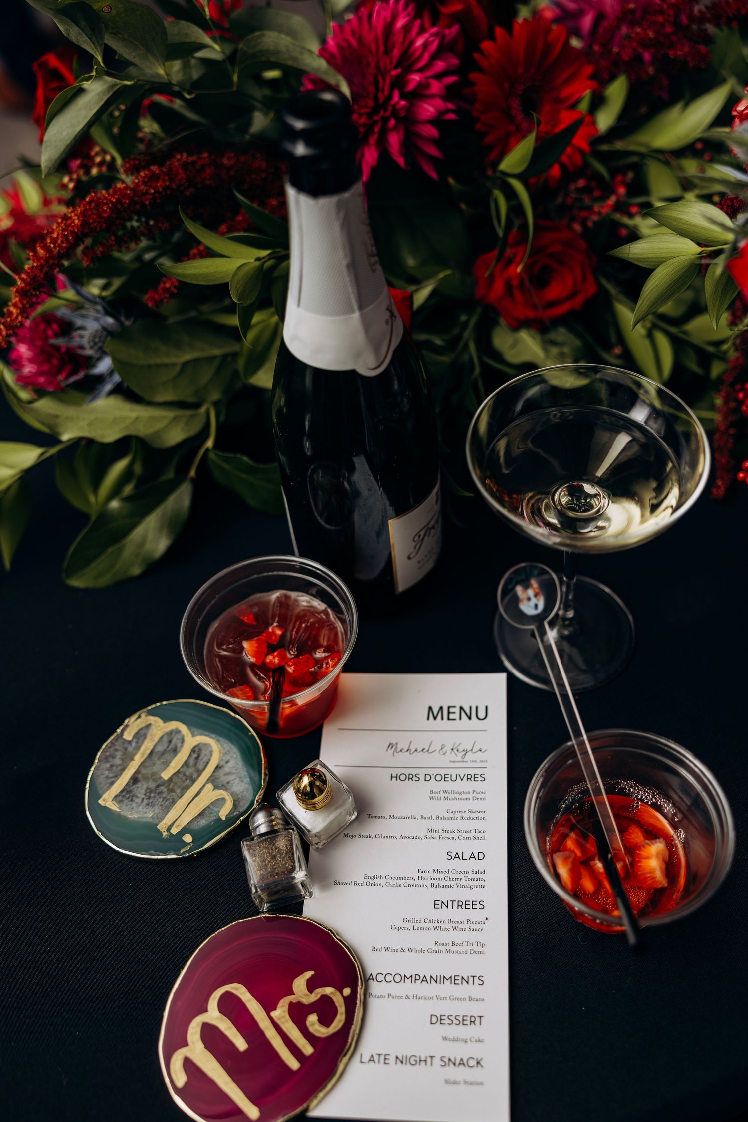 Elegant wedding table setting with champagne bottle, cocktails, Mr. and Mrs. tokens, menu card, and red floral centerpiece.