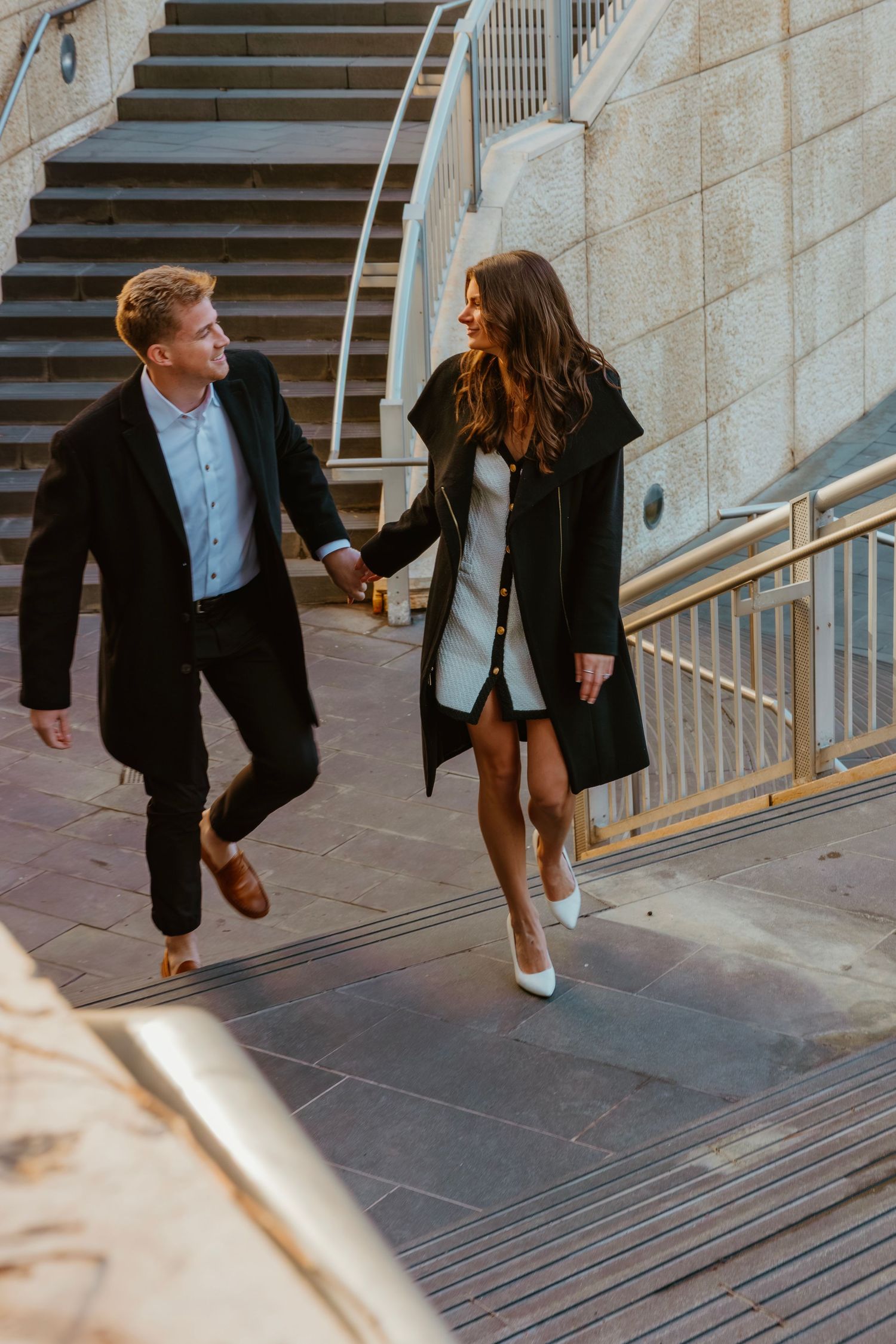 Smiling couple holding hands and climbing urban stairs together in elegant black outfits. Outdoor Chicago engagement photos.