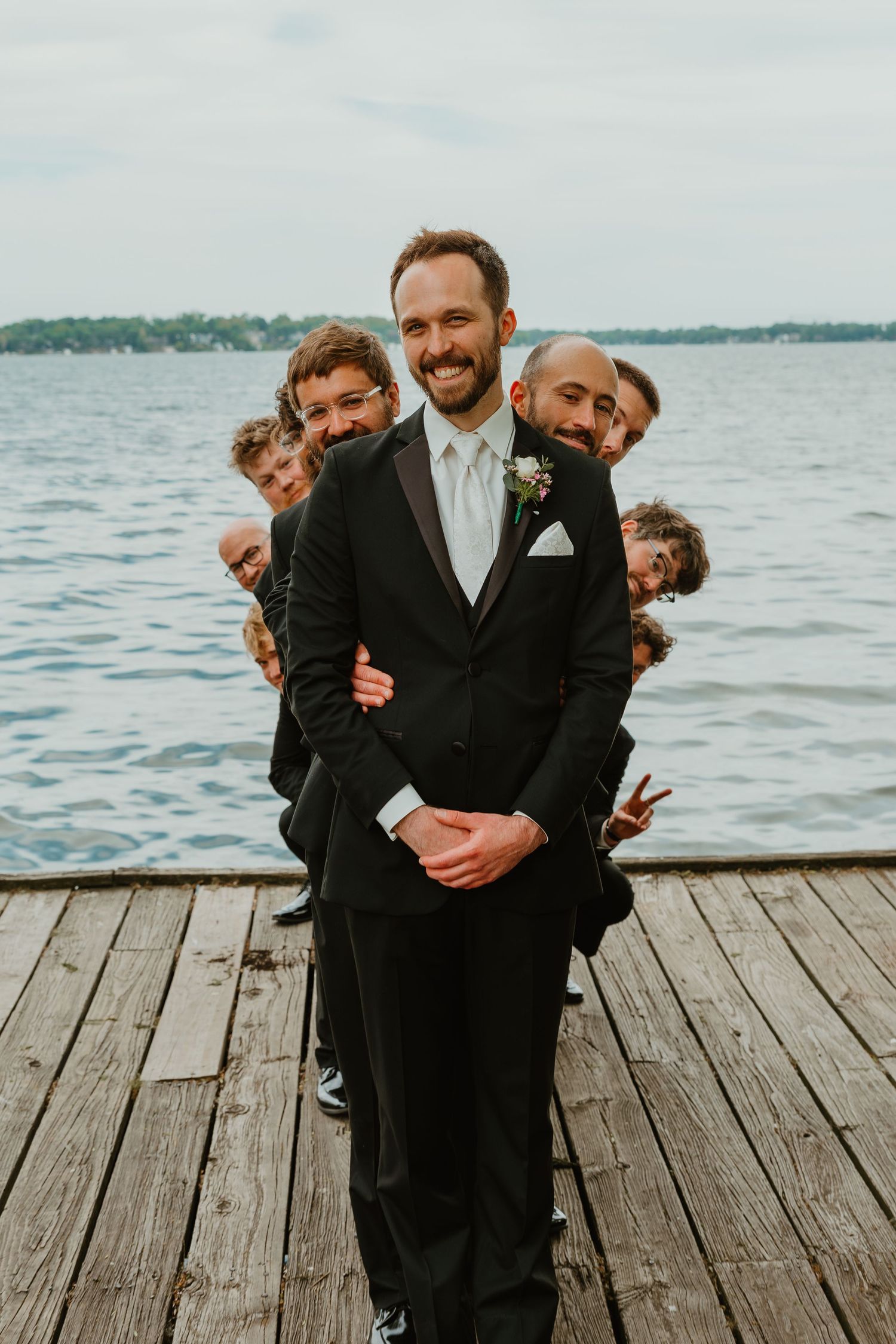 Smiling groom in black tuxedo poses on wooden dock as groomsmen playfully peek around him.