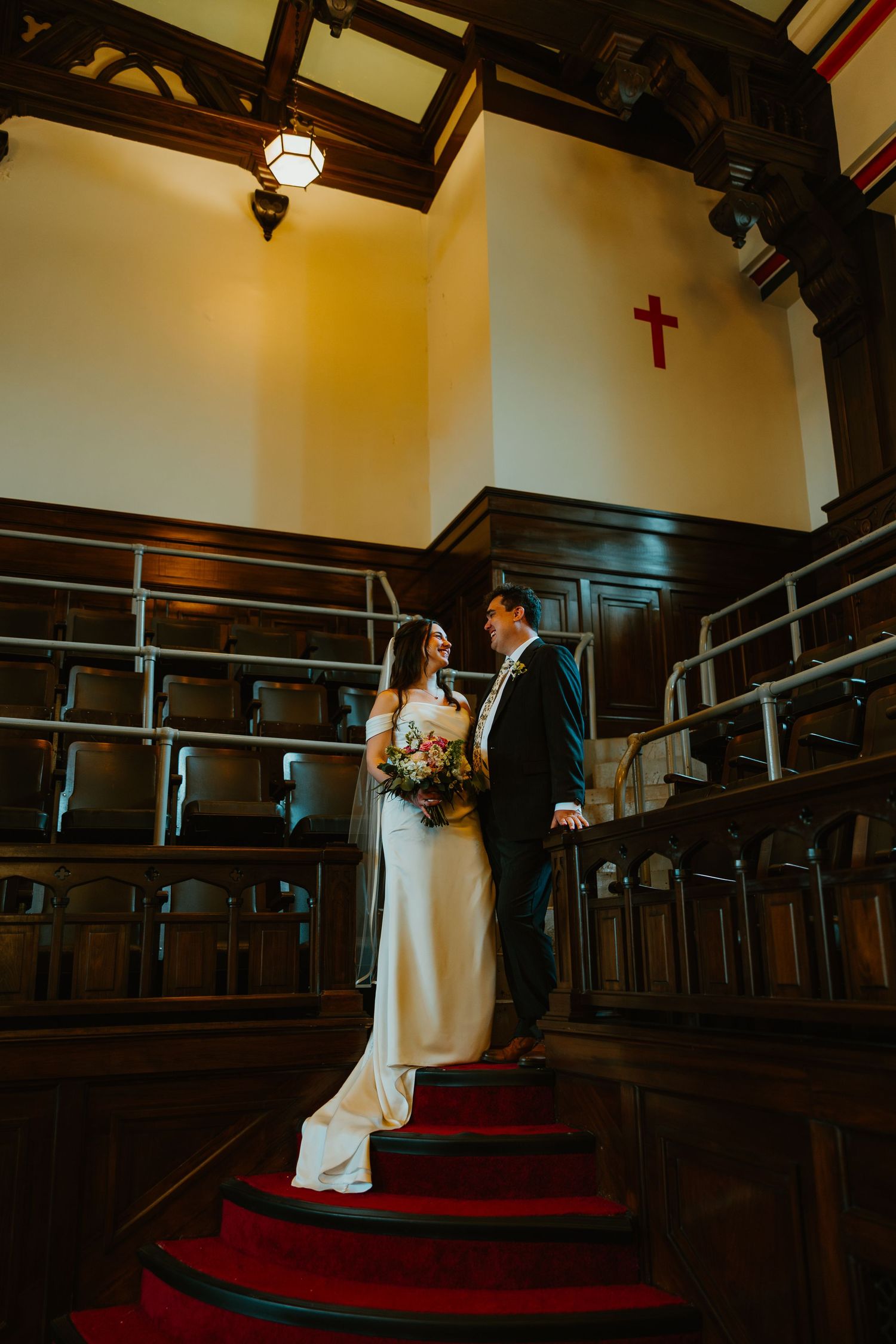 Bride and groom share an intimate moment on red-carpeted church stairs beneath a wooden cross.