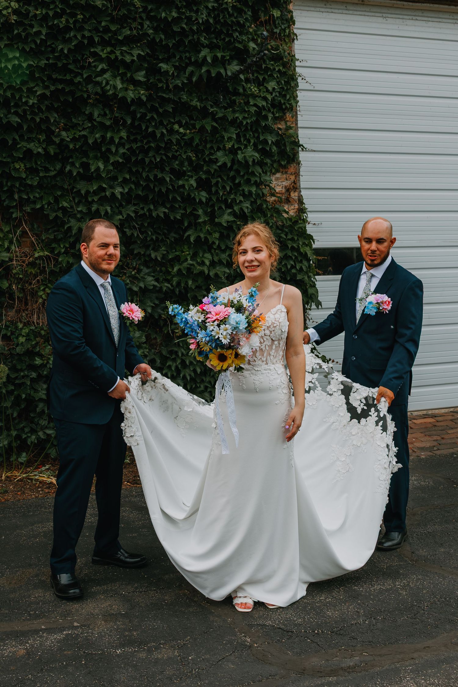 Bride in white gown holds colorful bouquet flanked by two groomsmen in navy suits holding her train outdoors. Chicagoland wedding venues.