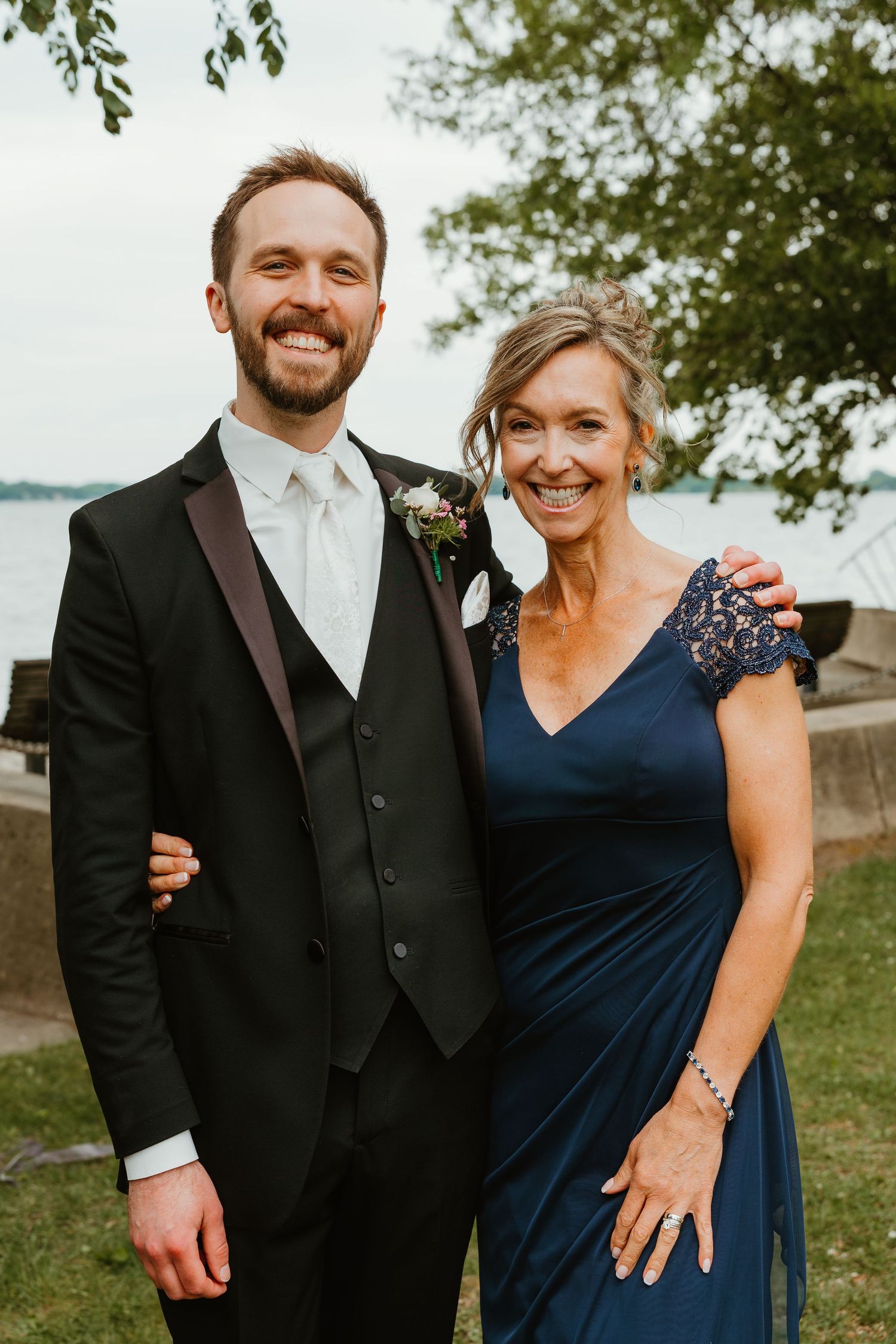 Smiling groom in black tuxedo with boutonniere posing with woman in navy lace dress by lakeside.