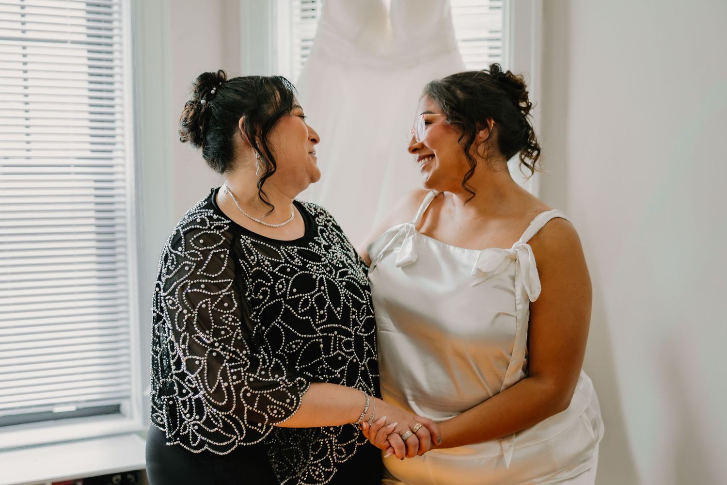 Bride and mother sharing a joyful moment while holding hands near a bright window. Chicagoland weddings.