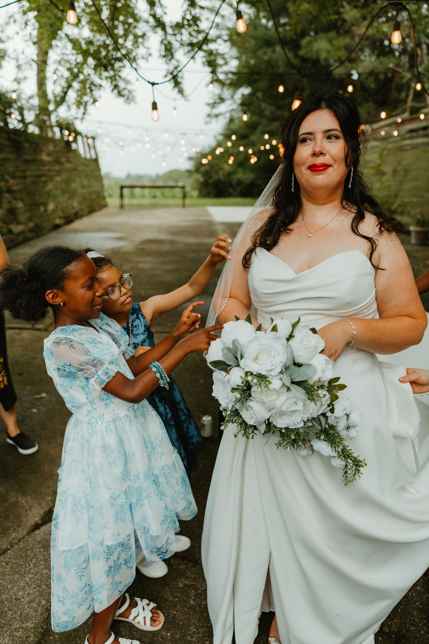 Bride in white strapless gown holds white rose bouquet while two young flower girls adjust her veil. Northwest Indiana wedding venues.