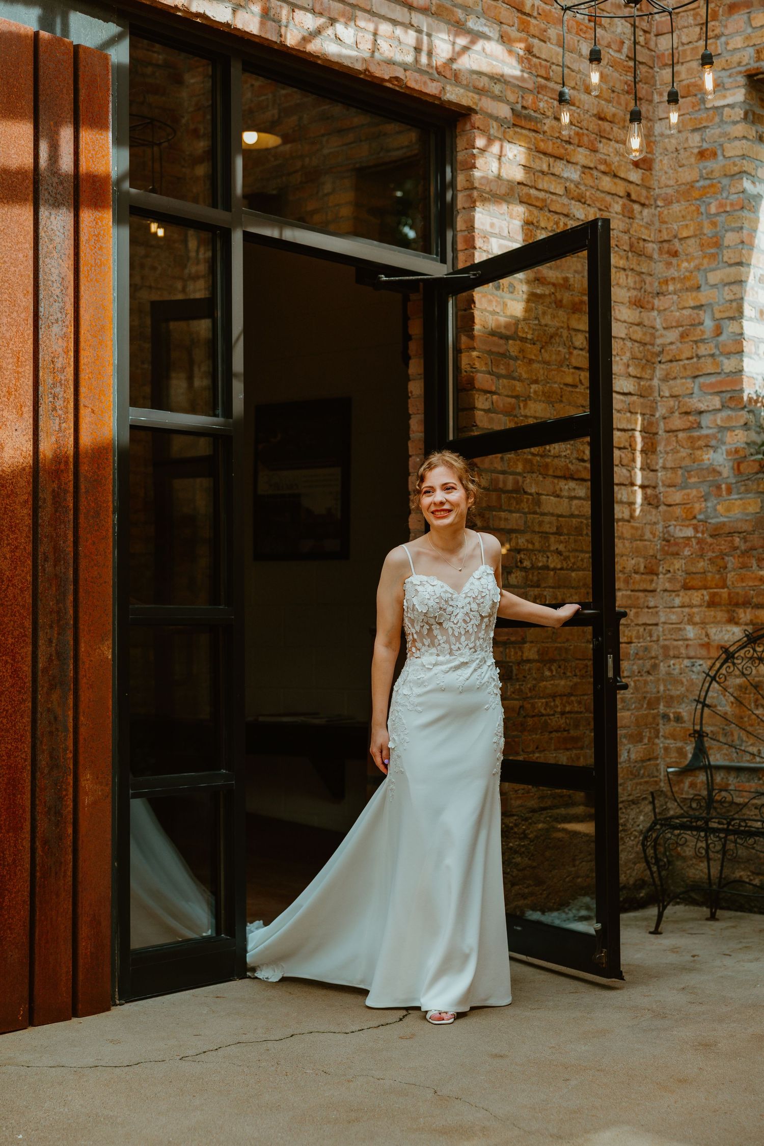 Smiling bride in a lace wedding gown standing in an open glass door of a brick building. Chicagoland wedding venues.