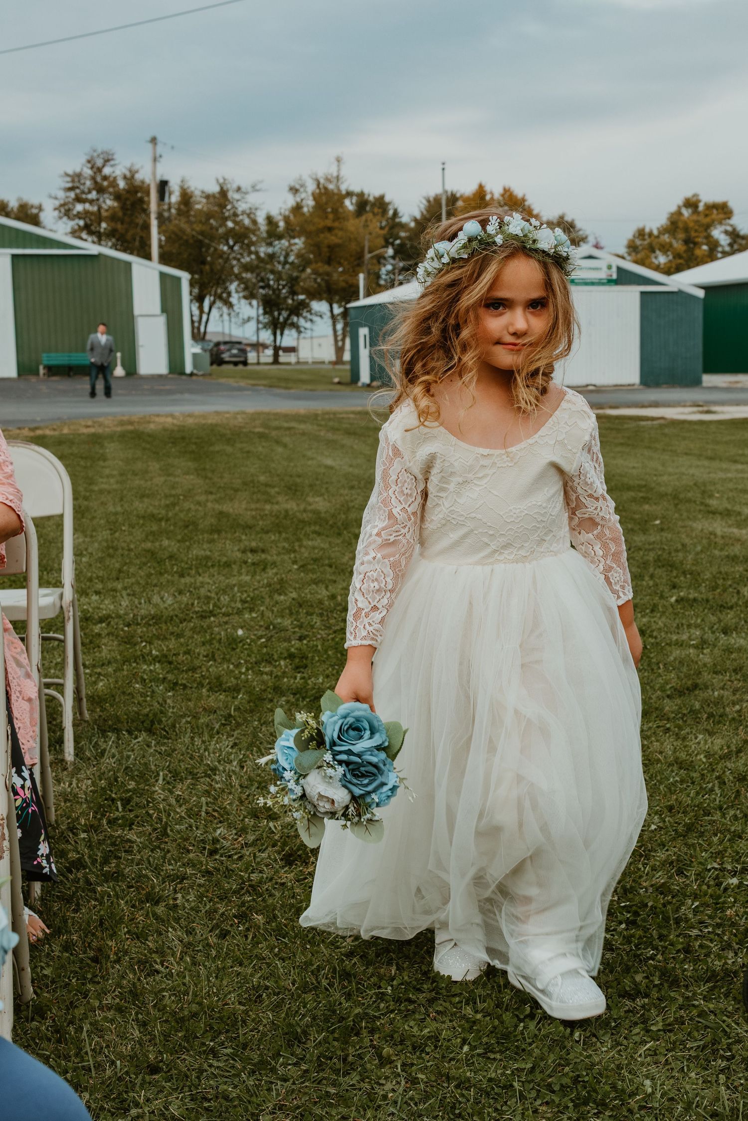 A young flower girl in a white lace dress and floral crown walks down the aisle holding blue roses. Northwest Indiana wedding venues.