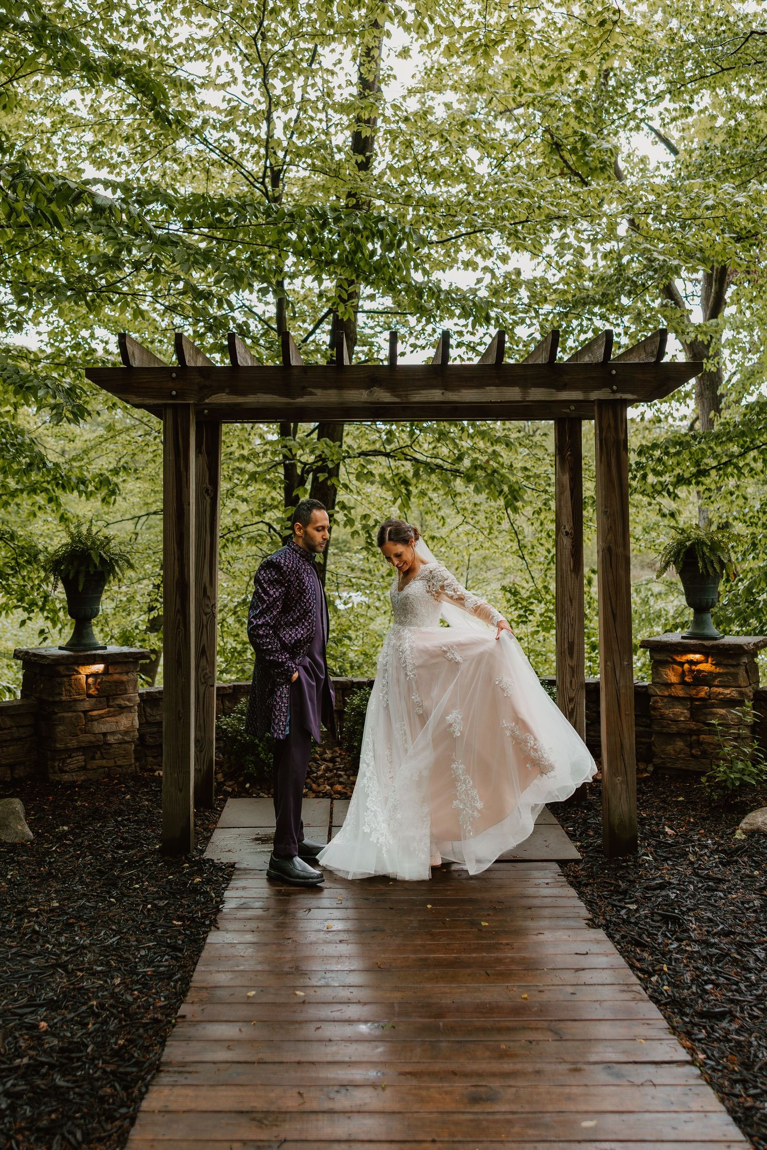 Bride and groom standing under a wooden pergola arch surrounded by lush green trees.