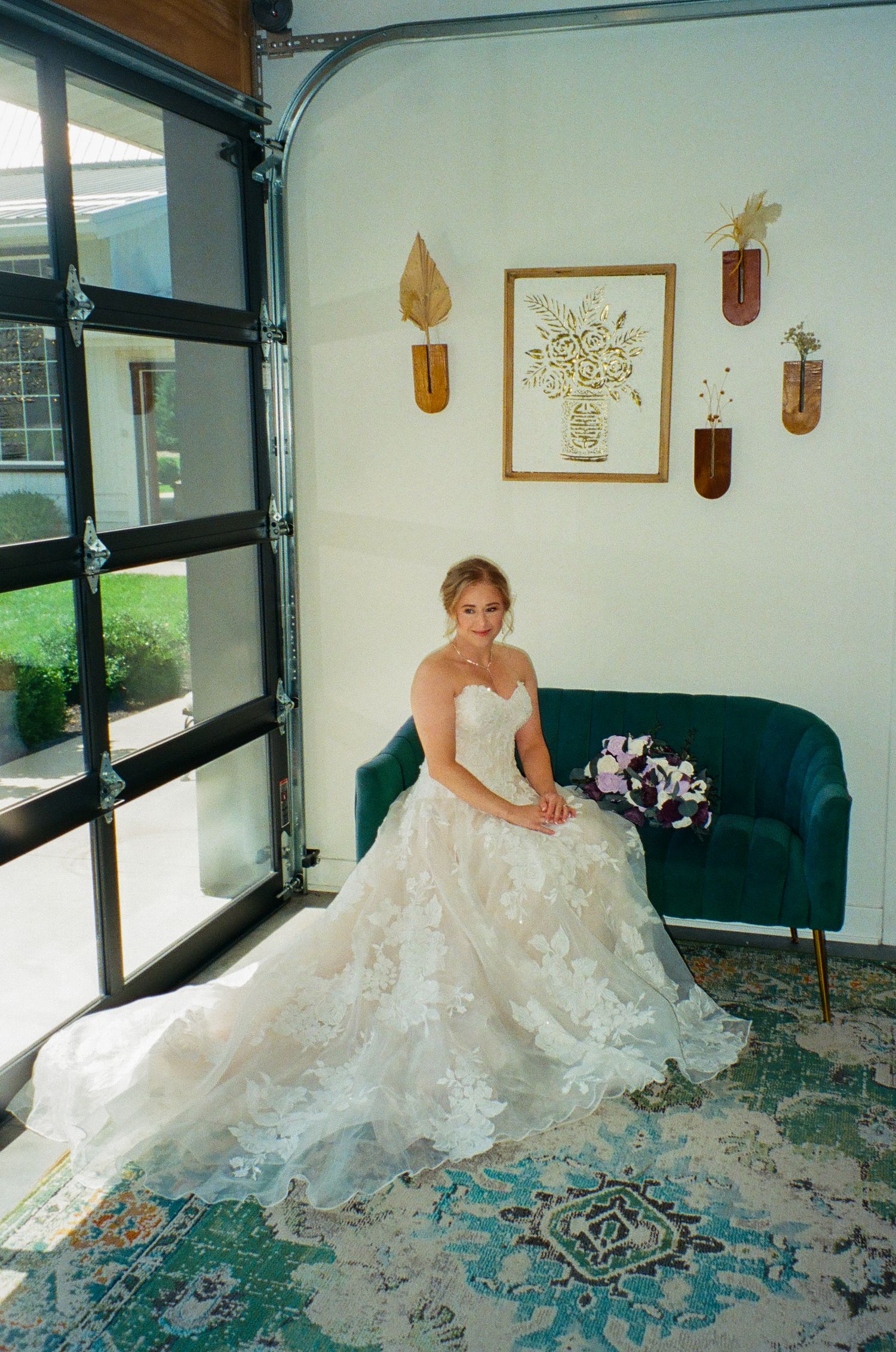 Bride in strapless lace ballgown seated on teal velvet sofa holding purple bouquet beside large window in elegant venue.