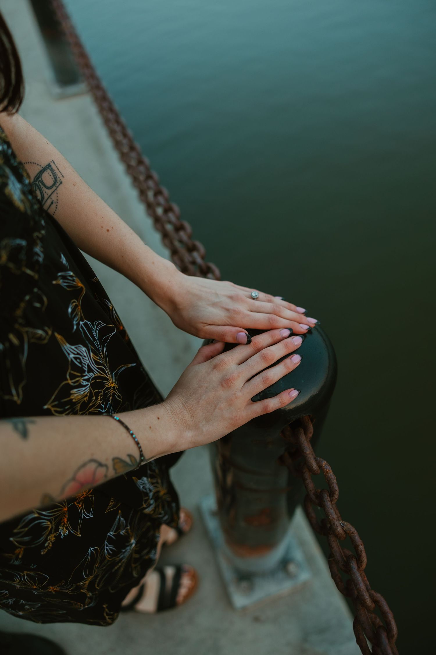 Close-up of tattooed woman's hands resting on a dock post near water, wearing an engagement ring. Chicagoland engagement photos.