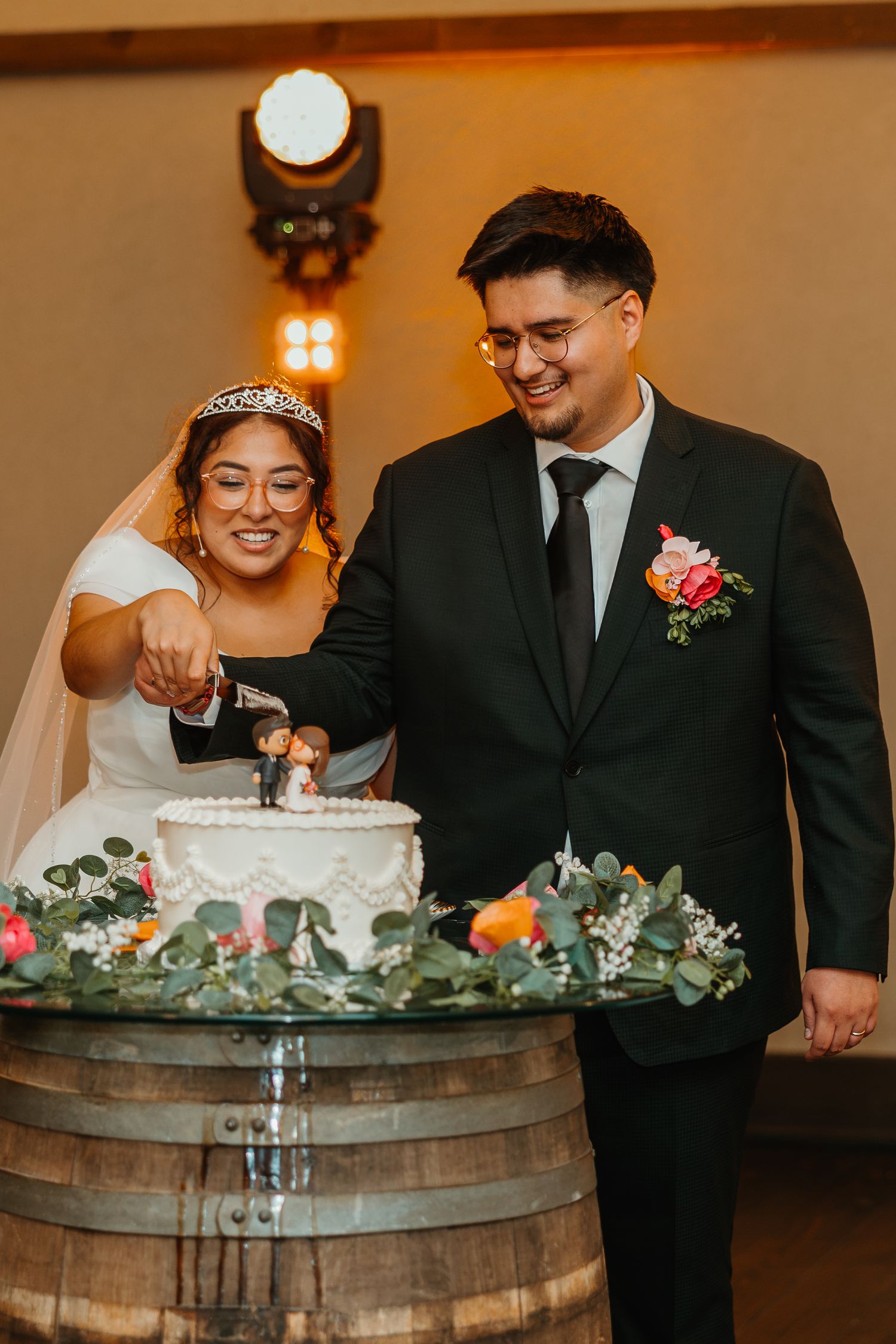 Happy newlyweds cutting white wedding cake displayed on a rustic barrel surrounded by colorful floral garland. Chicagoland wedding venues.