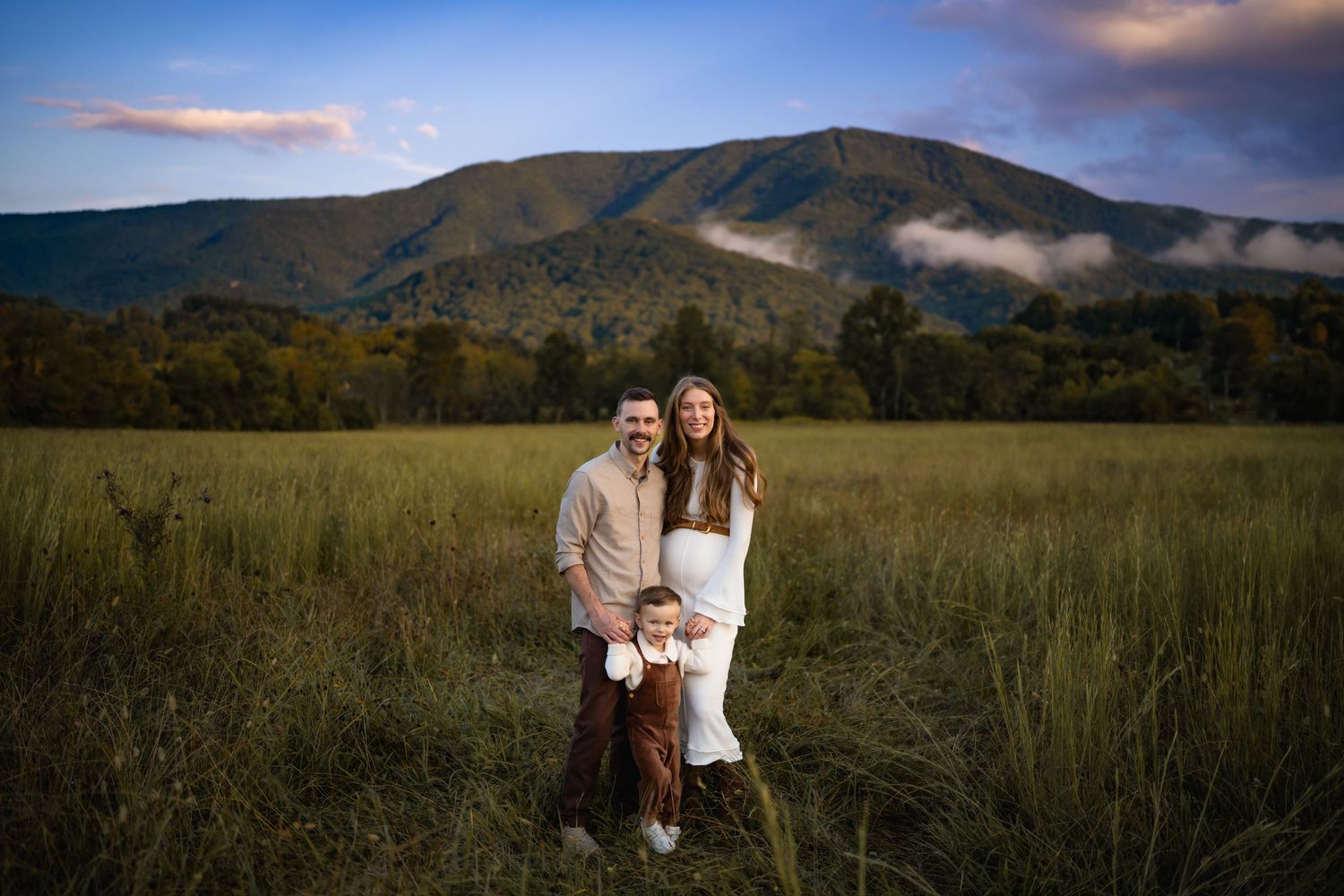 Family of three posing in tall grass field with mountain and clouds in background at sunset.