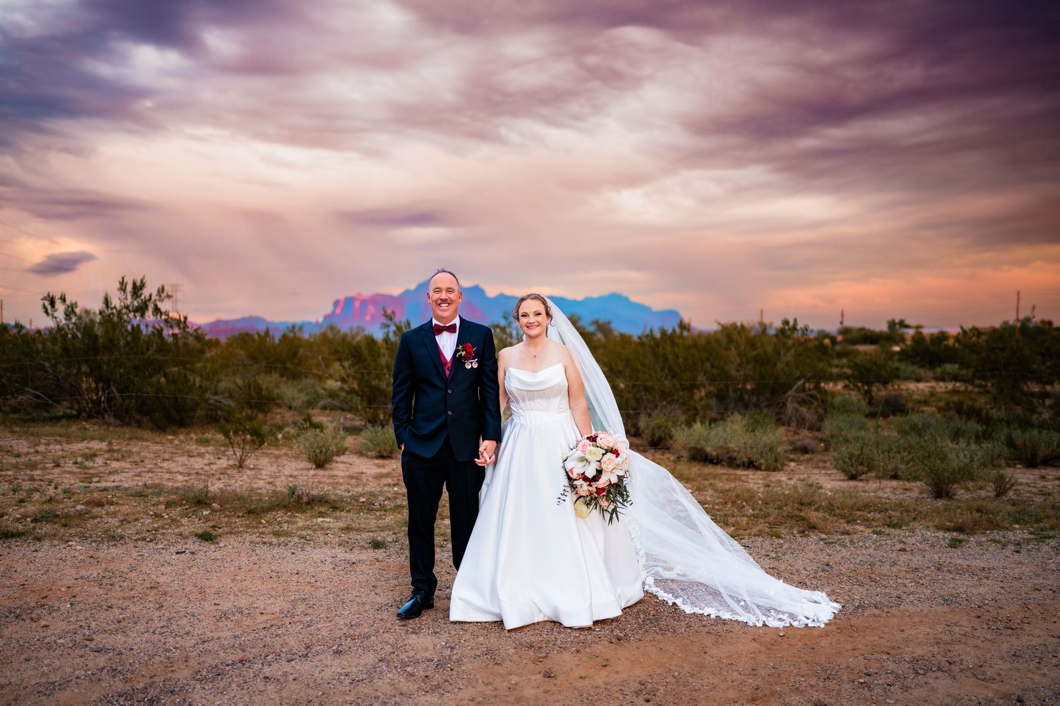 Bride and groom pose in desert landscape at sunset with purple mountains and dramatic sky backdrop.
