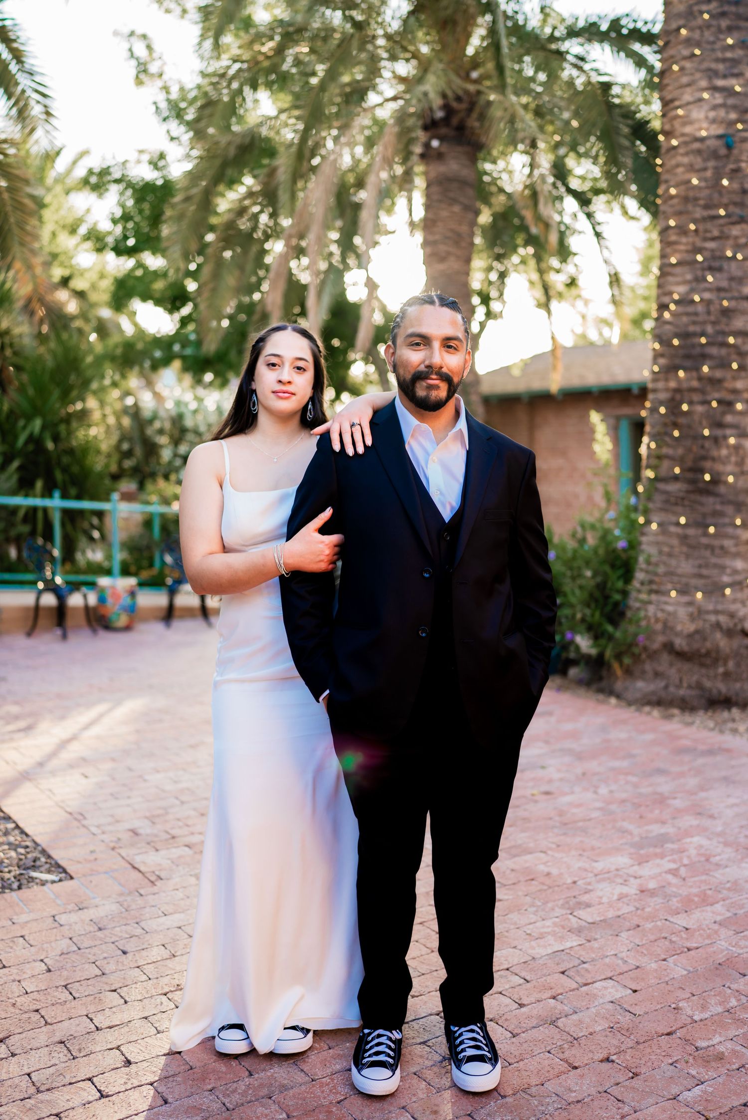 Couple posing outdoors in formal attire, woman in white gown and man in black suit, surrounded by palm trees.