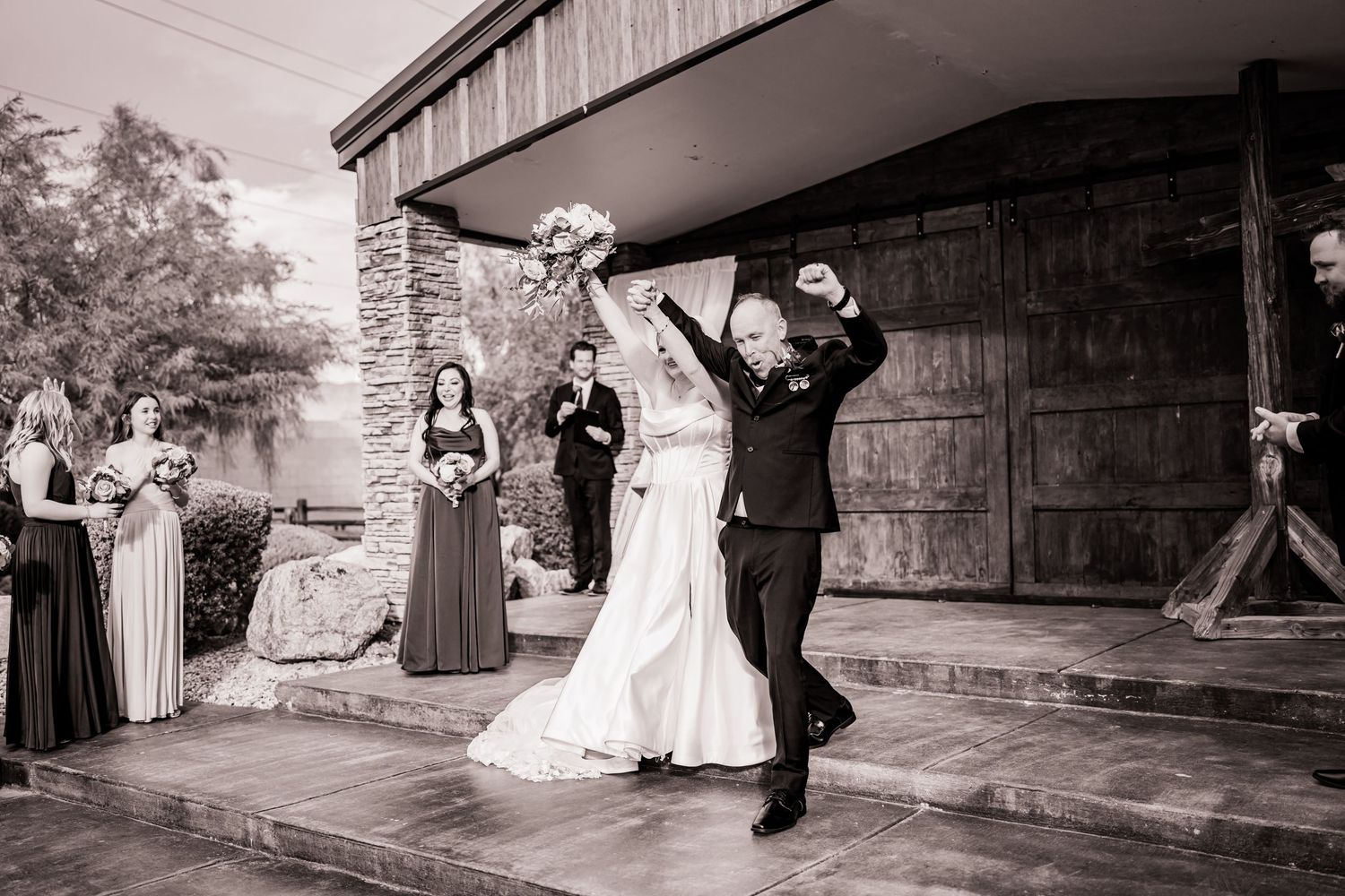 Black and white photo of joyful newlyweds celebrating after wedding ceremony with bridesmaids and groomsmen in background.