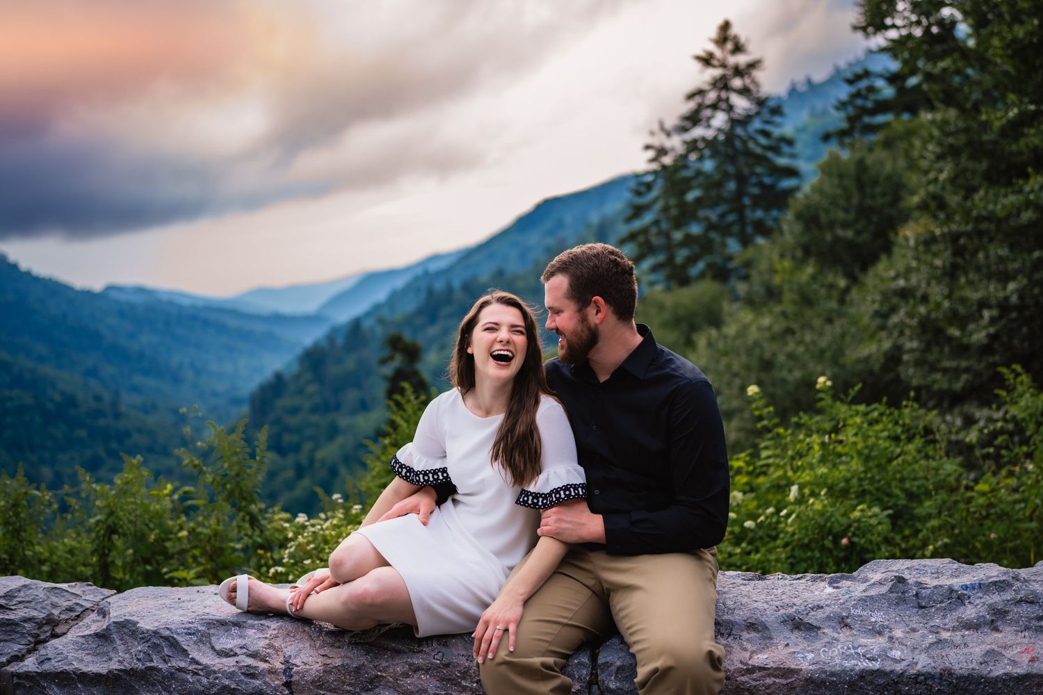 Couple laughing together on stone wall with dramatic mountain sunset backdrop.