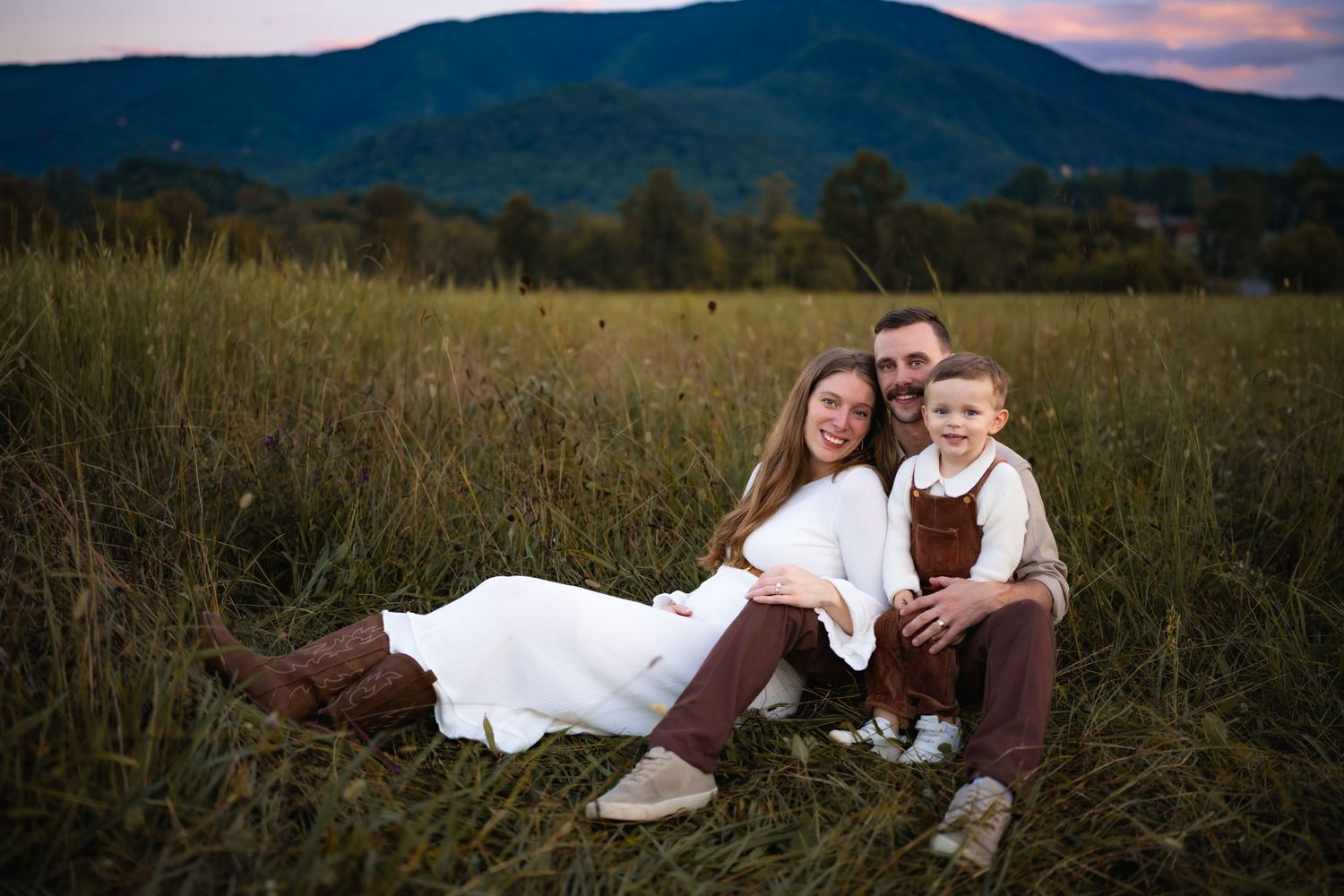 Family of three sitting together in a golden grass field with purple mountain sunset backdrop.