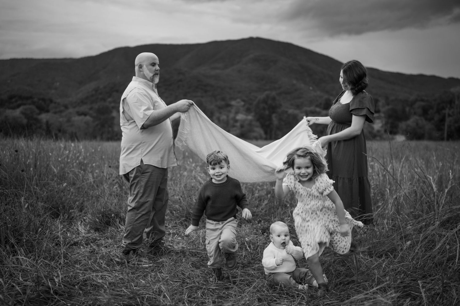 Black and white family photo in a field with mountains, parents holding a blanket over children playing outdoors.