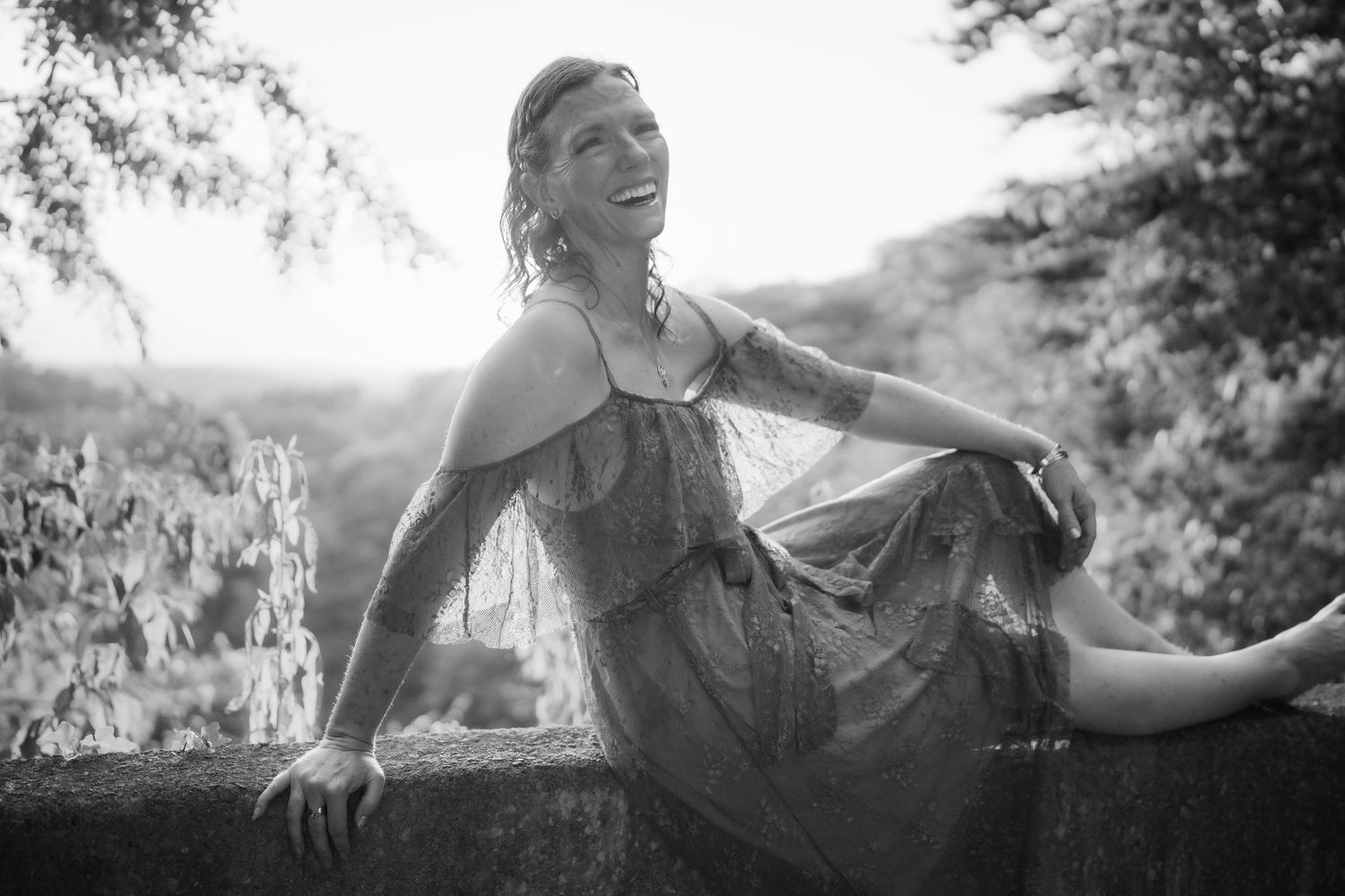 Black and white photo of a smiling woman in a flowy dress seated on a stone wall outdoors.