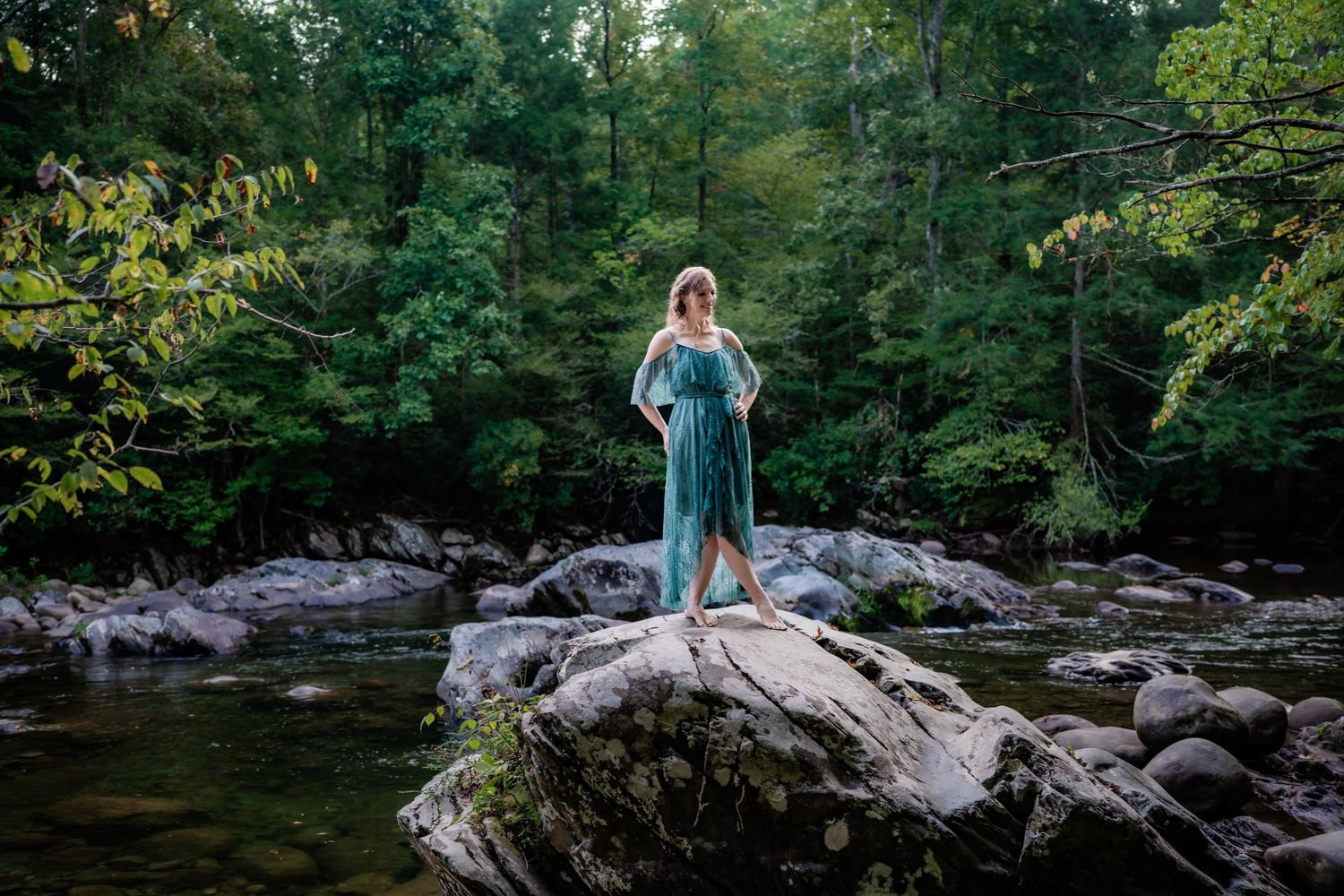Woman in teal dress standing on river rocks surrounded by lush forest.