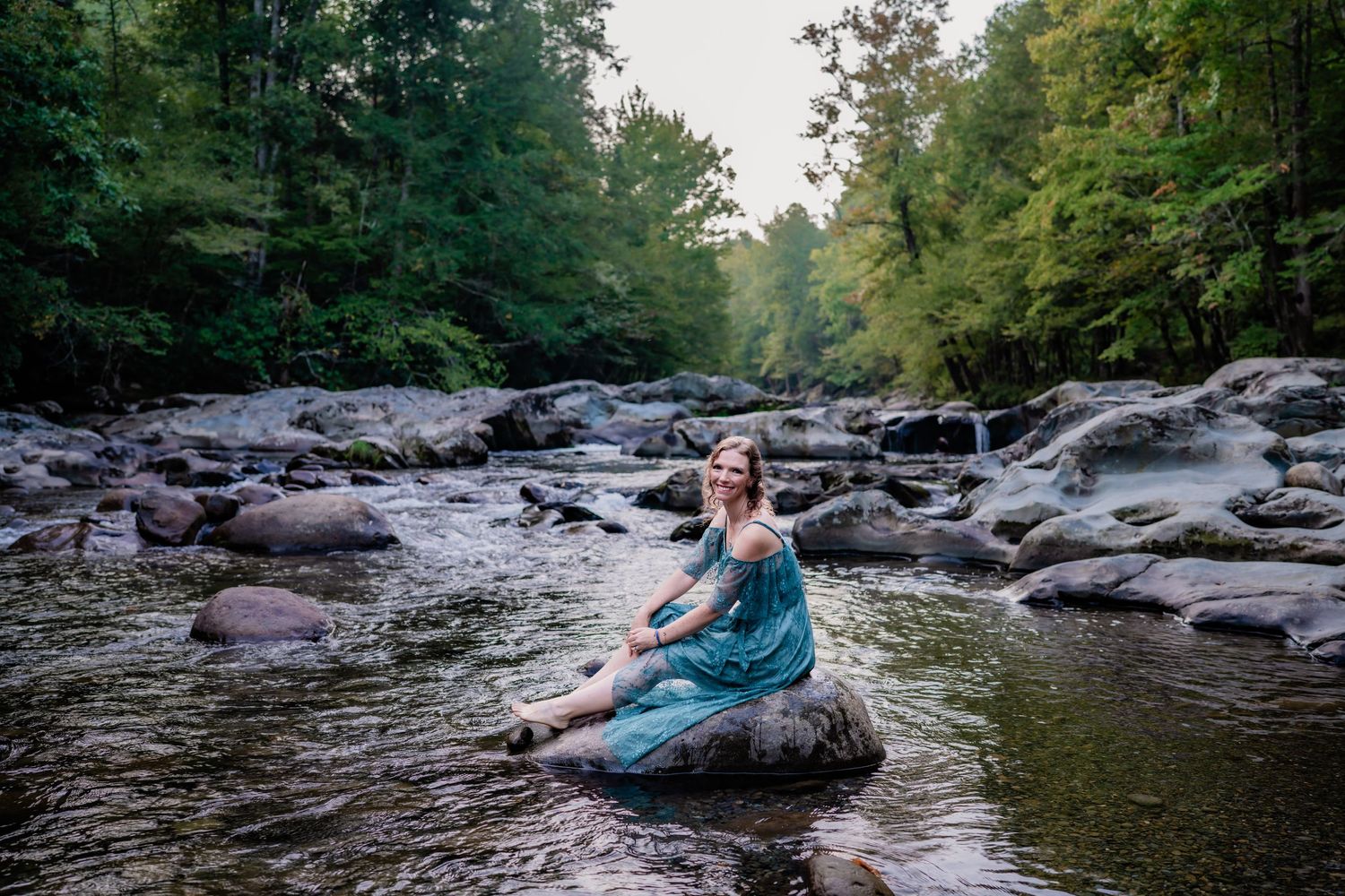 Woman in teal dress sitting on rocks in a peaceful forest river.