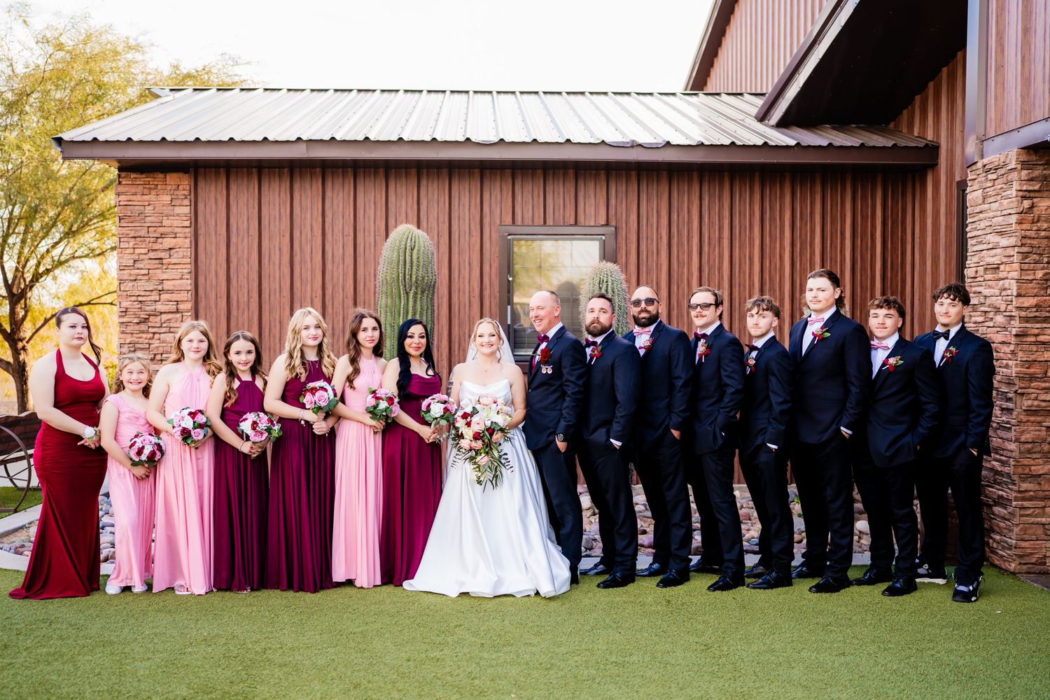 Wedding party poses outdoors, bridesmaids in pink and burgundy gowns, groomsmen in navy suits, bride in white gown.