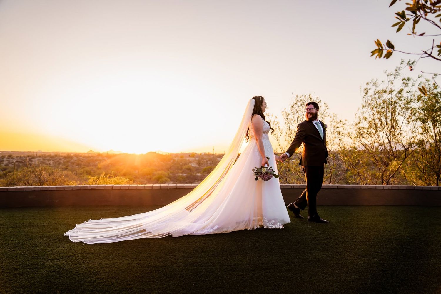 Bride and groom holding hands at sunset on a rooftop overlooking a city skyline, bride wearing a long white gown and veil.