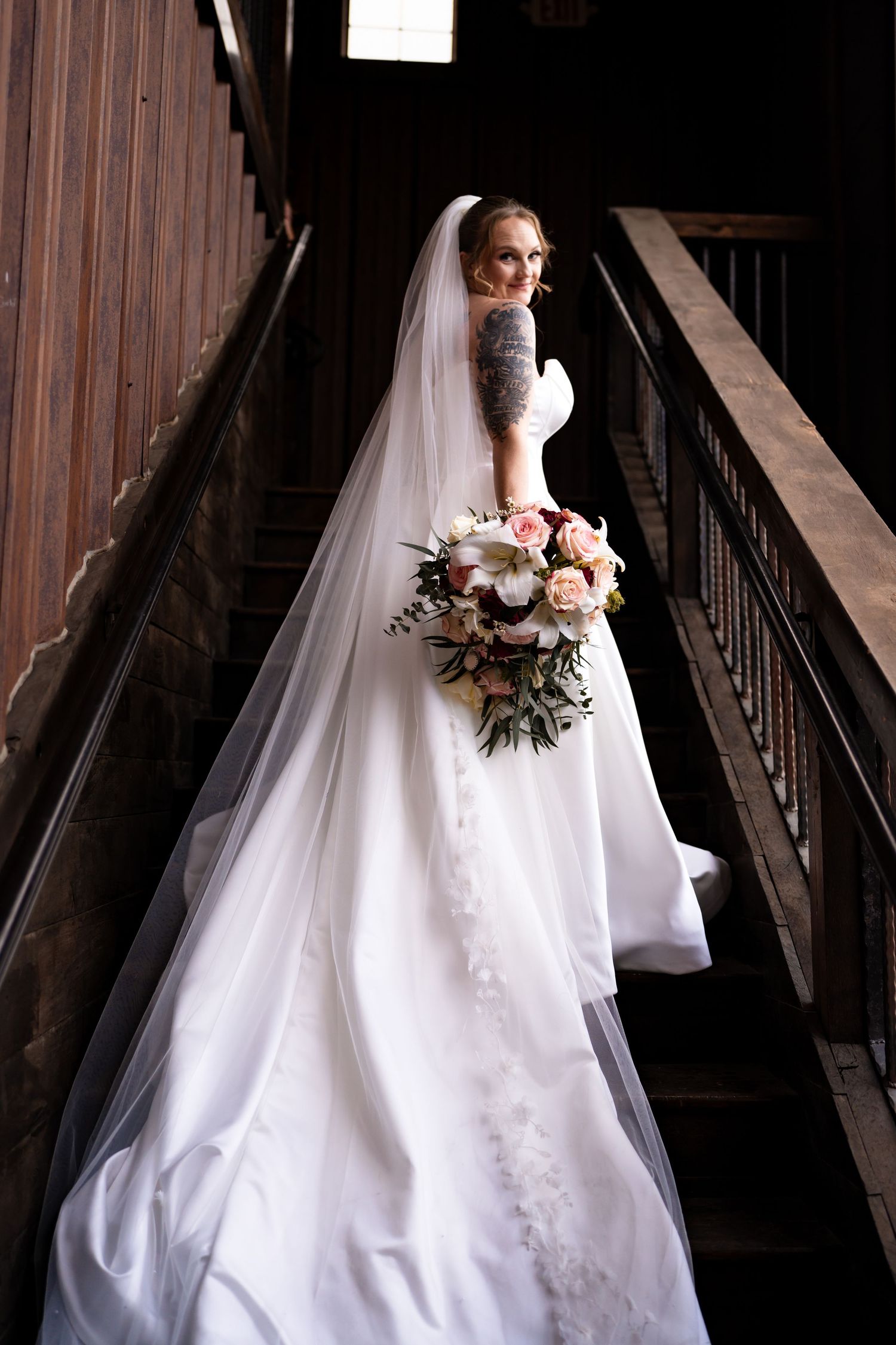 Bride in white gown and veil holds bouquet on wooden staircase, looking back over shoulder.