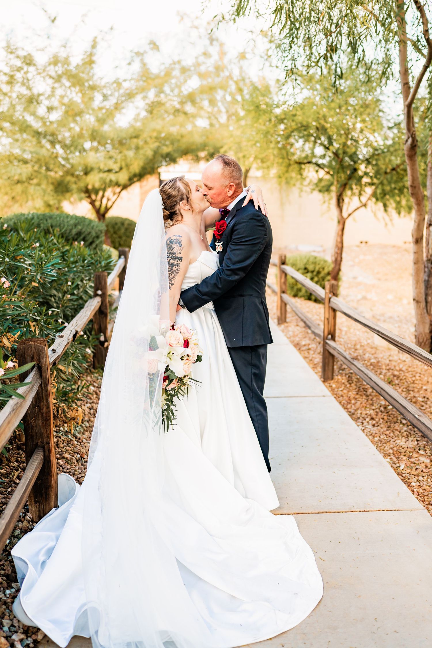 Bride and groom share a romantic kiss on a garden pathway, bride wearing a flowing white gown with veil and bouquet.