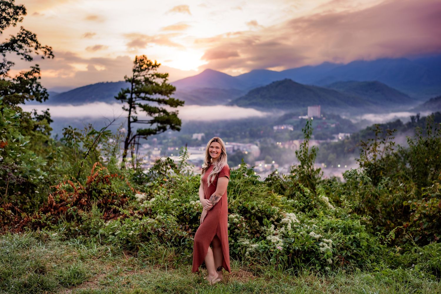 Woman in red dress poses on misty mountain overlook at sunset with foggy valley and blue ridgeline behind her.
