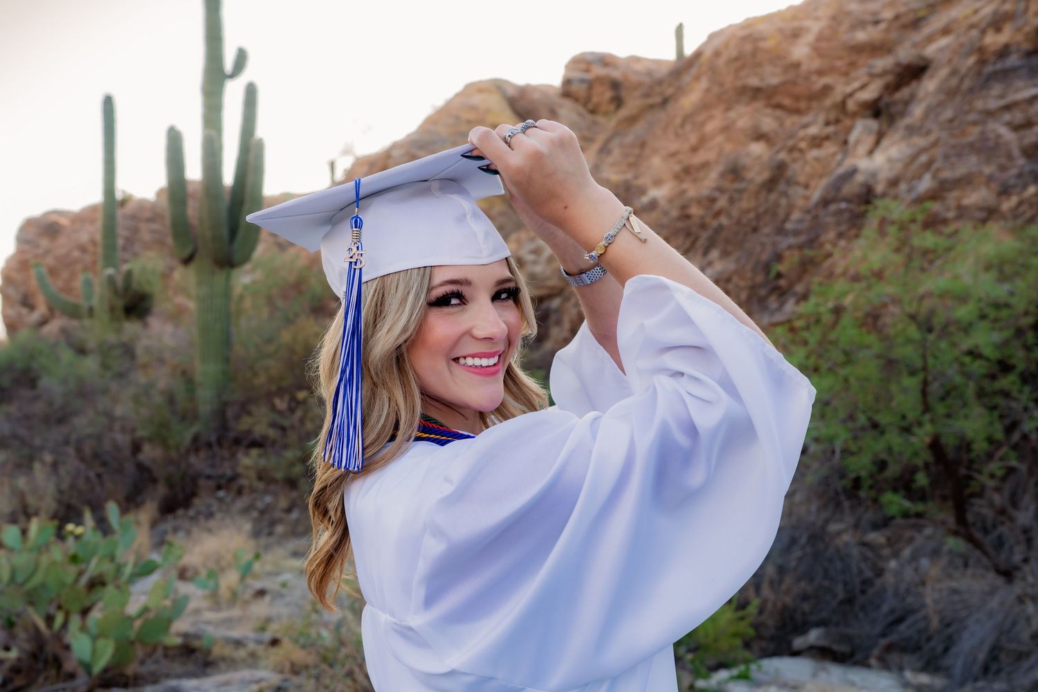 A smiling graduate in white cap and gown poses outdoors in a desert landscape with saguaro cacti and rock formations.