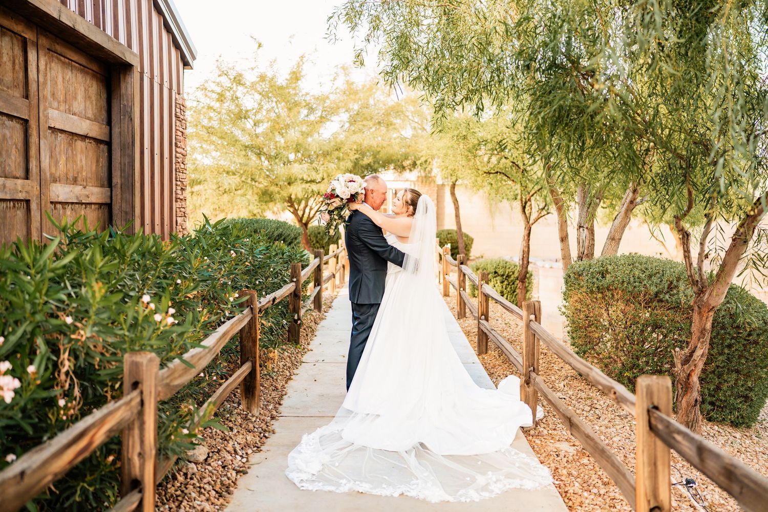 Bride and groom share a romantic kiss on a wooden bridge pathway surrounded by lush green trees at an outdoor wedding venue.