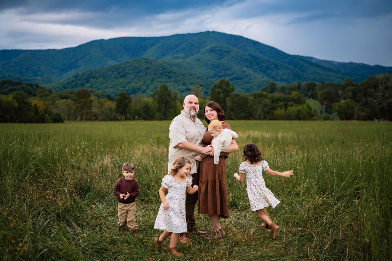 Family of five posing in a lush green meadow with blue mountains and dramatic sky behind them.