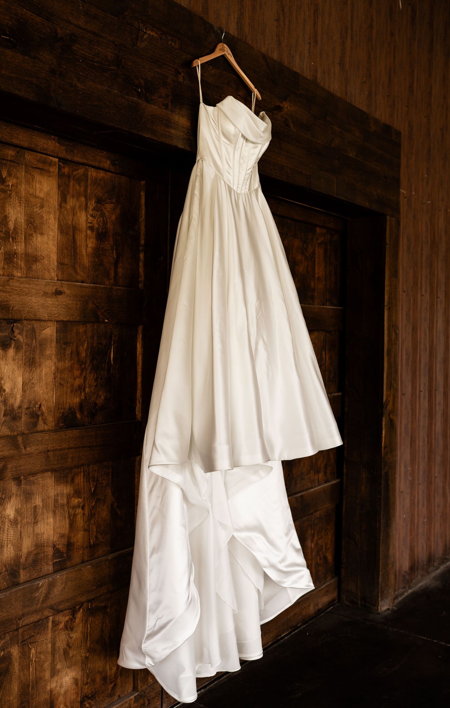 Elegant white satin wedding gown with sweetheart neckline hanging on wooden hanger against rustic dark wood wall.