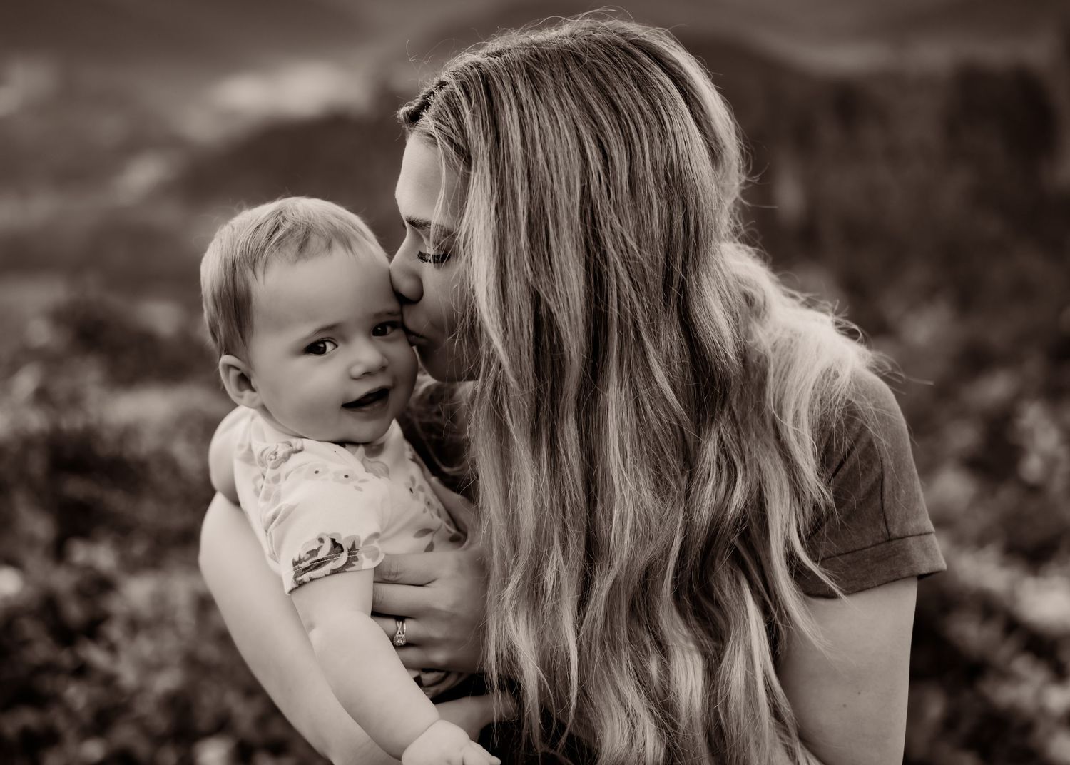 Black and white photo of a mother kissing her smiling baby outdoors in a tender, loving moment.