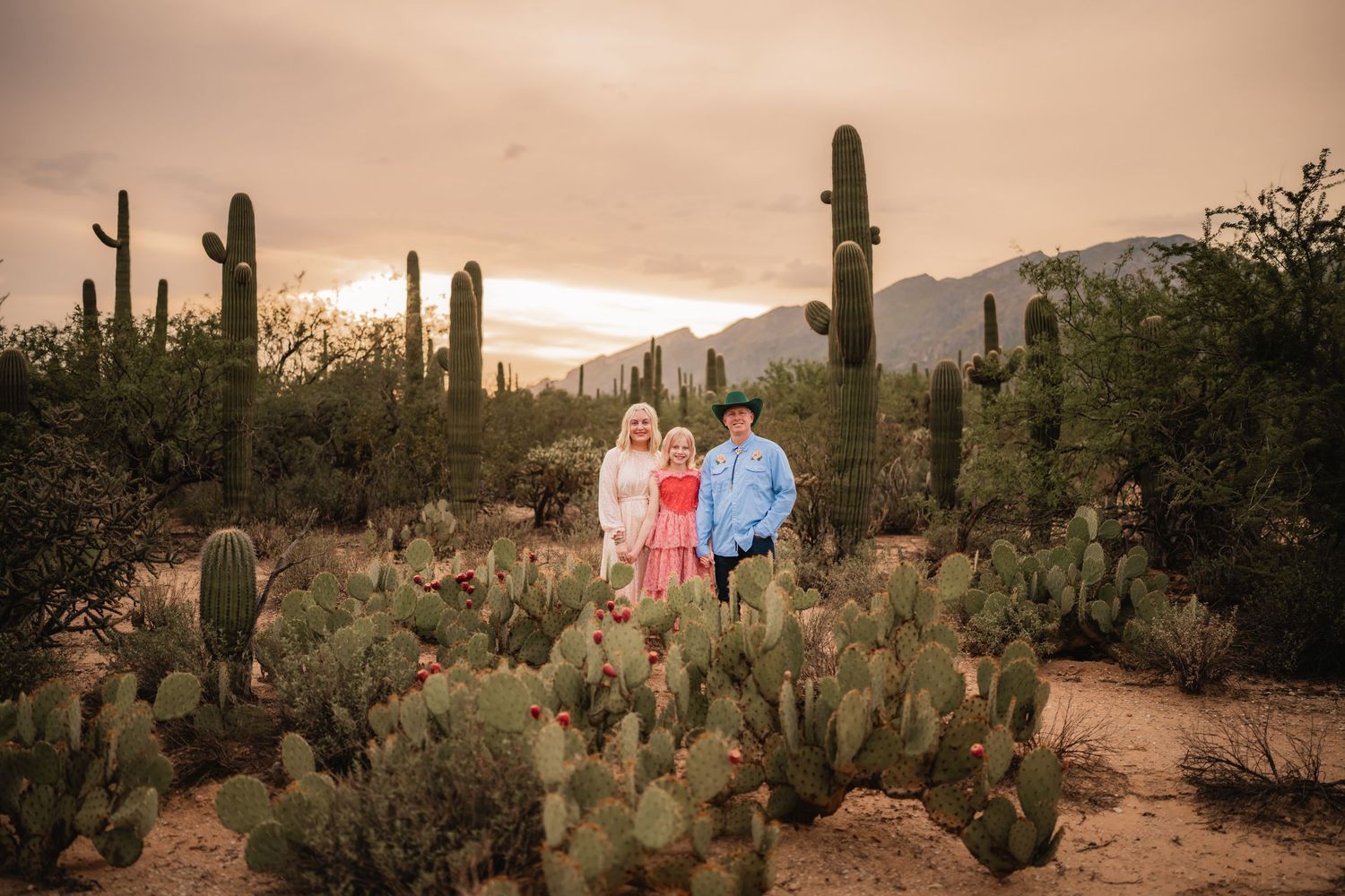 Family of three posing among saguaro cacti and prickly pear in Arizona desert at golden sunset.