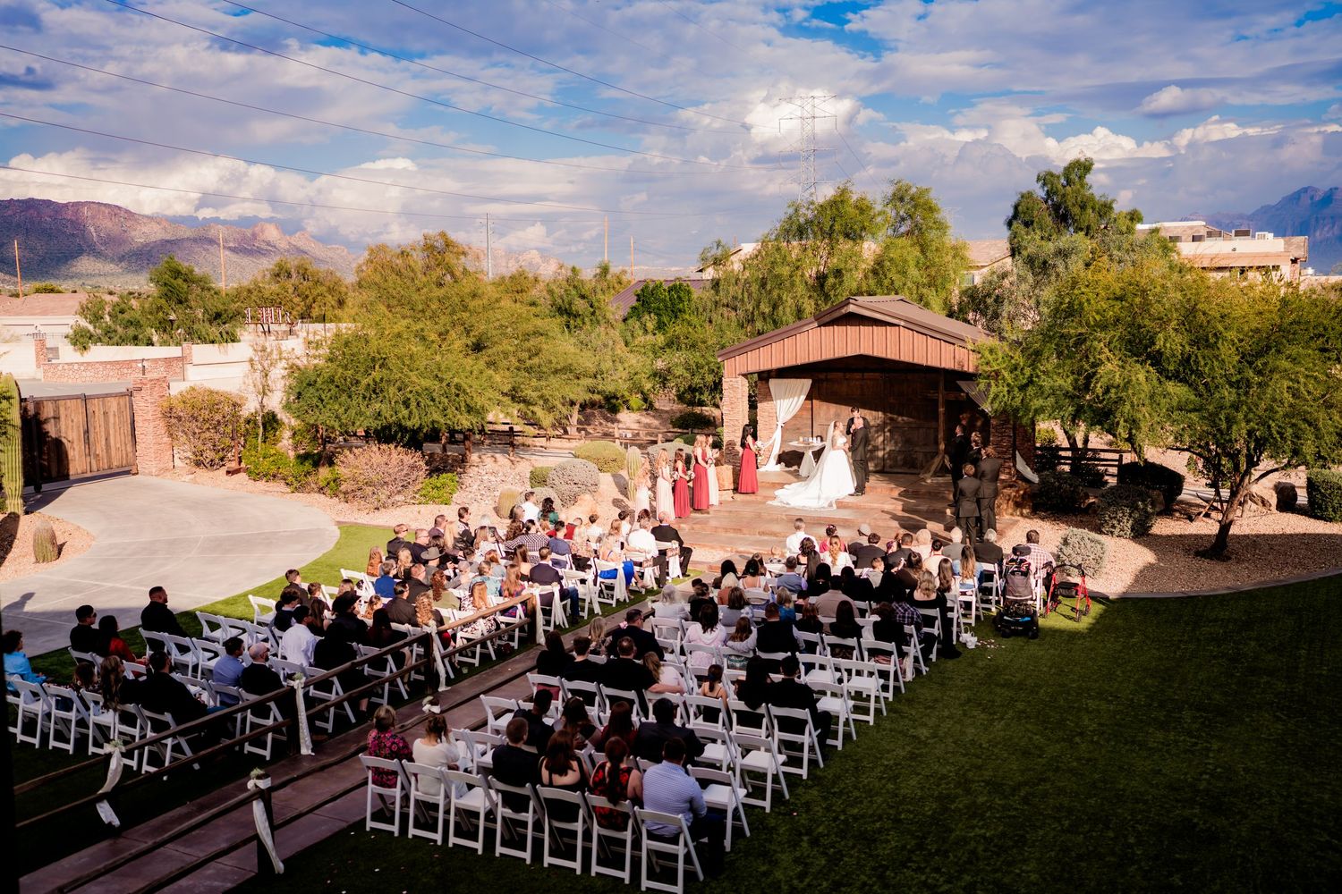 Outdoor wedding ceremony with guests seated on white chairs facing a rustic stage, set against a scenic desert landscape.