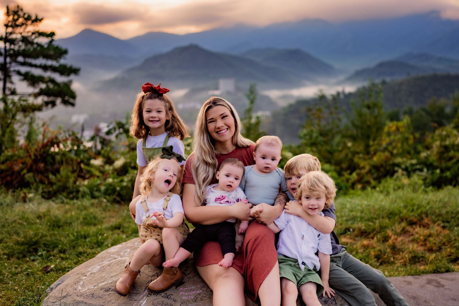 Woman sits on rock with six young children against misty mountain sunset backdrop.