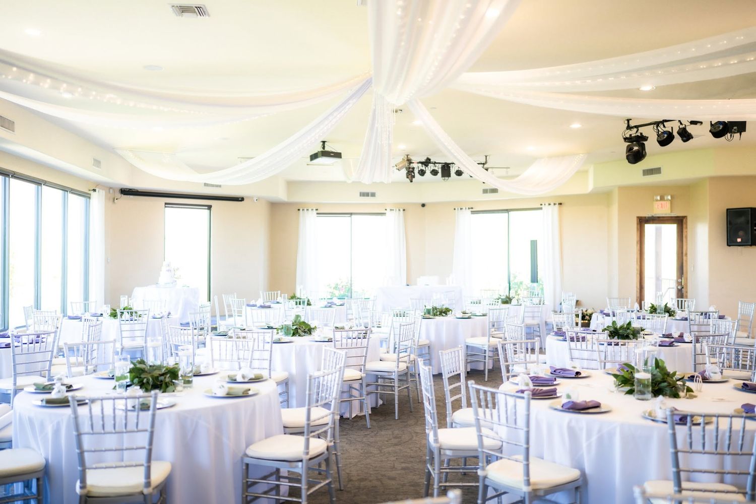 Elegant wedding reception hall with white draped ceilings, round tables, white chiavari chairs, and floral centerpieces.