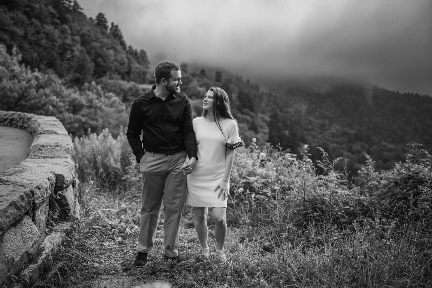 Black and white photo of a couple sharing a romantic moment on a scenic mountain overlook surrounded by lush trees.