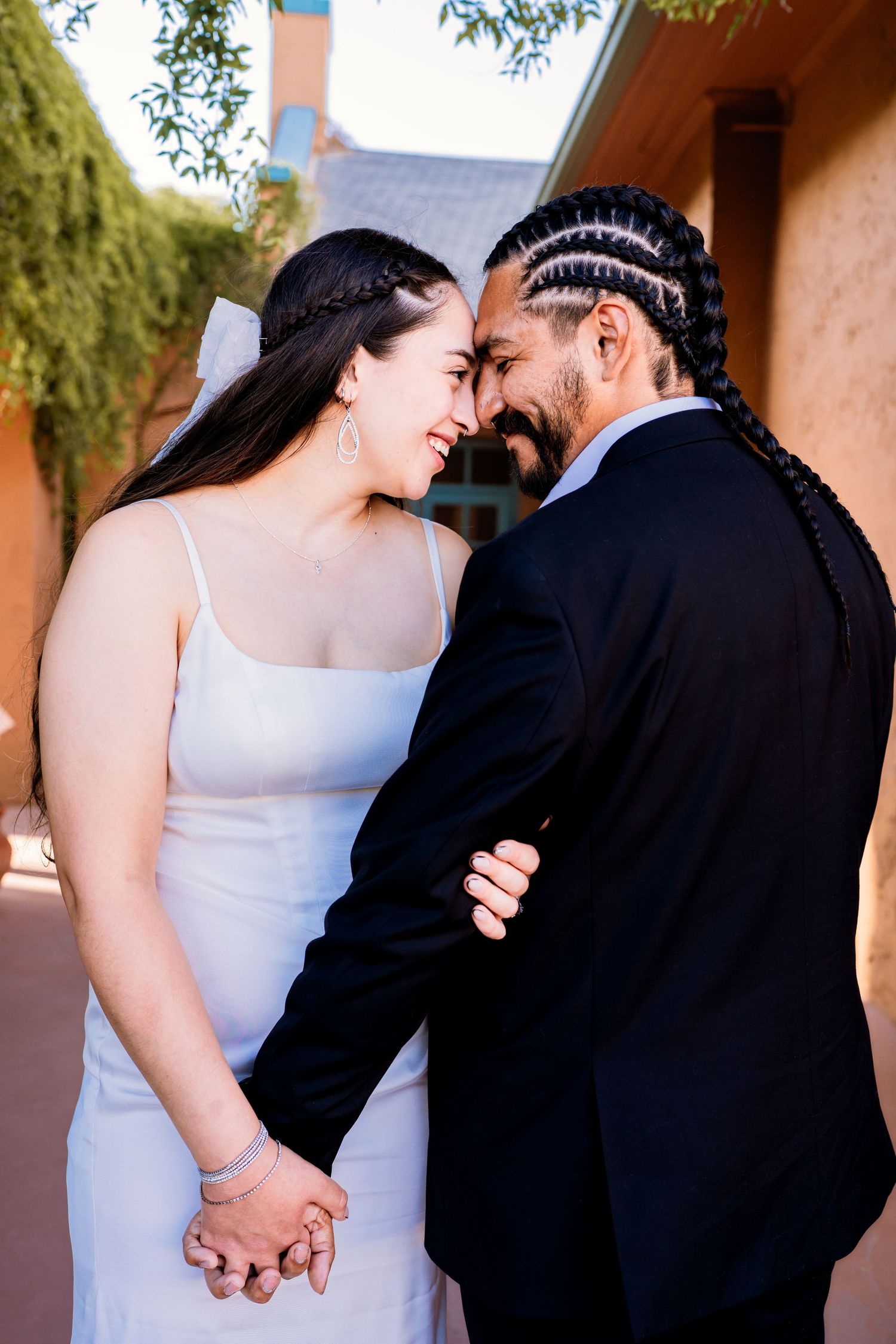 A bride and groom in wedding attire smile forehead-to-forehead while holding hands outdoors.