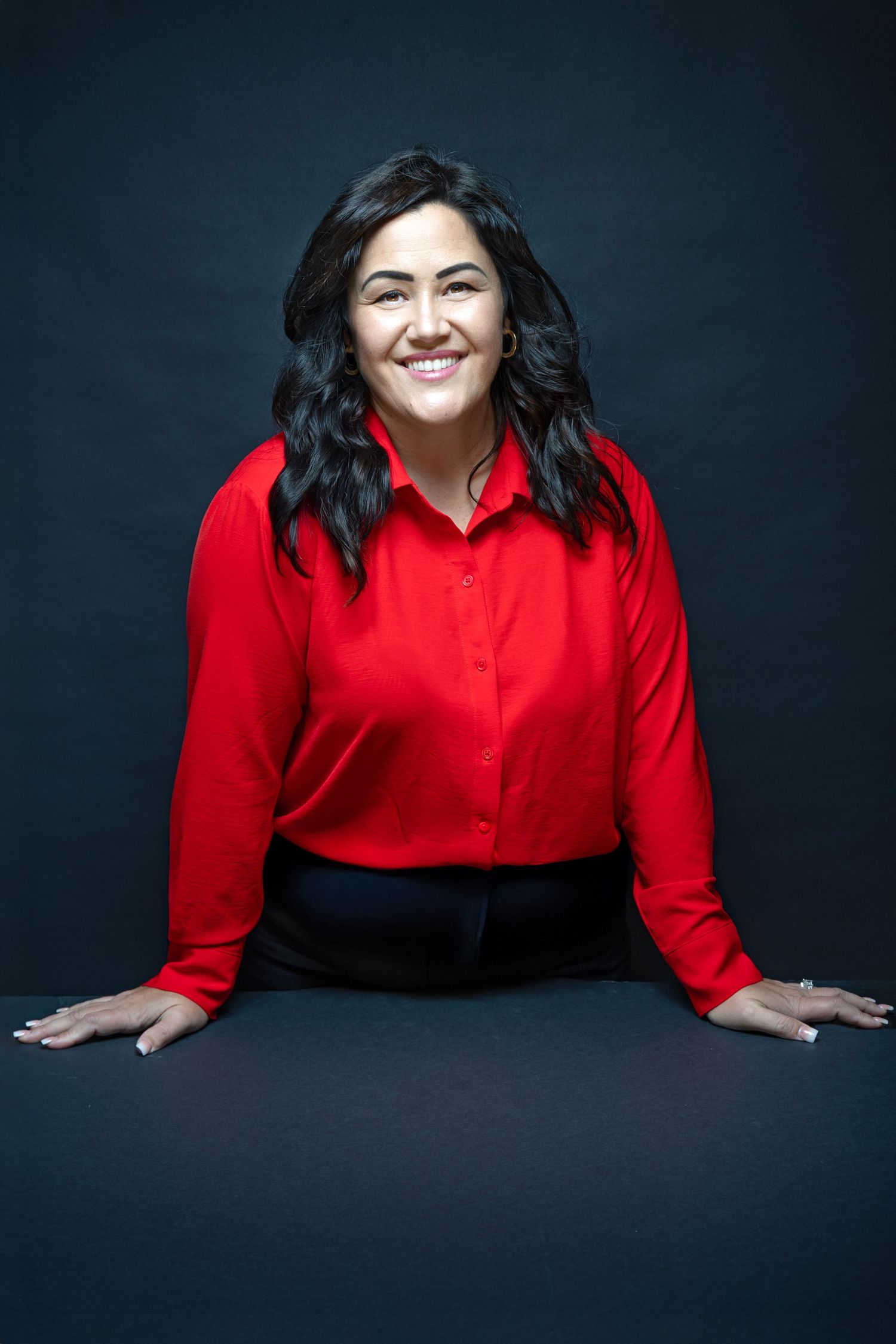 Professional woman in red blouse smiling confidently while leaning on a table against dark background.