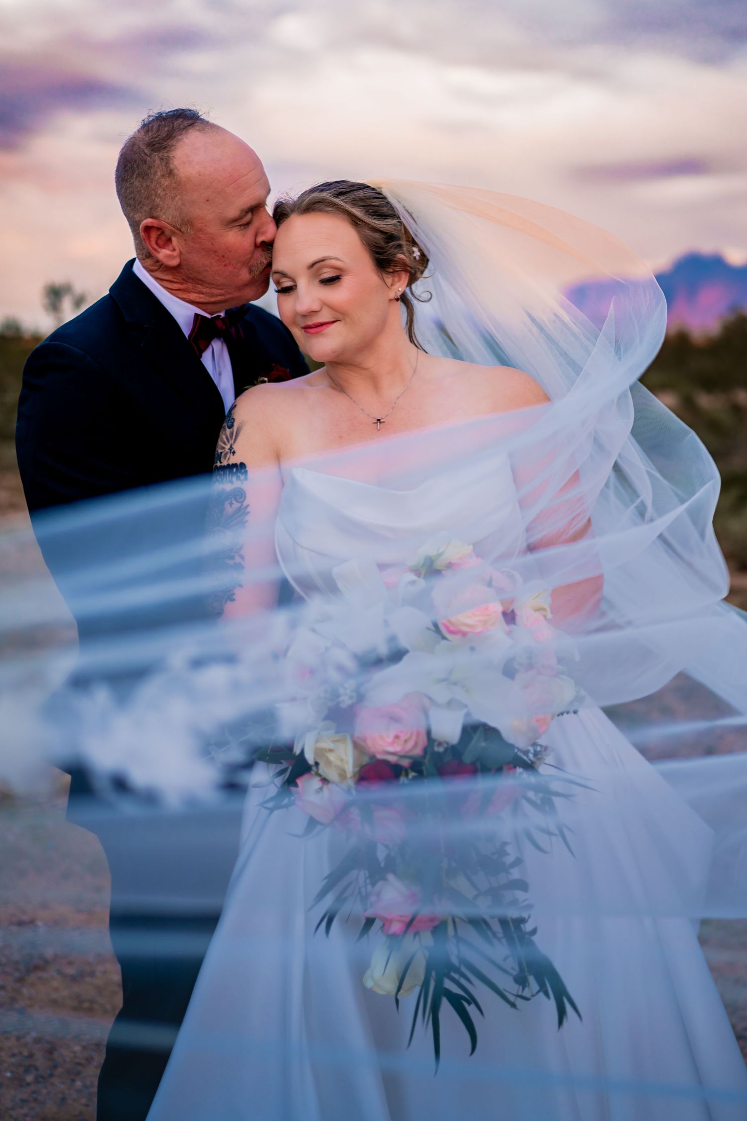 Bride and groom share a romantic kiss at sunset, with flowing veil and pink rose bouquet against a purple sky.