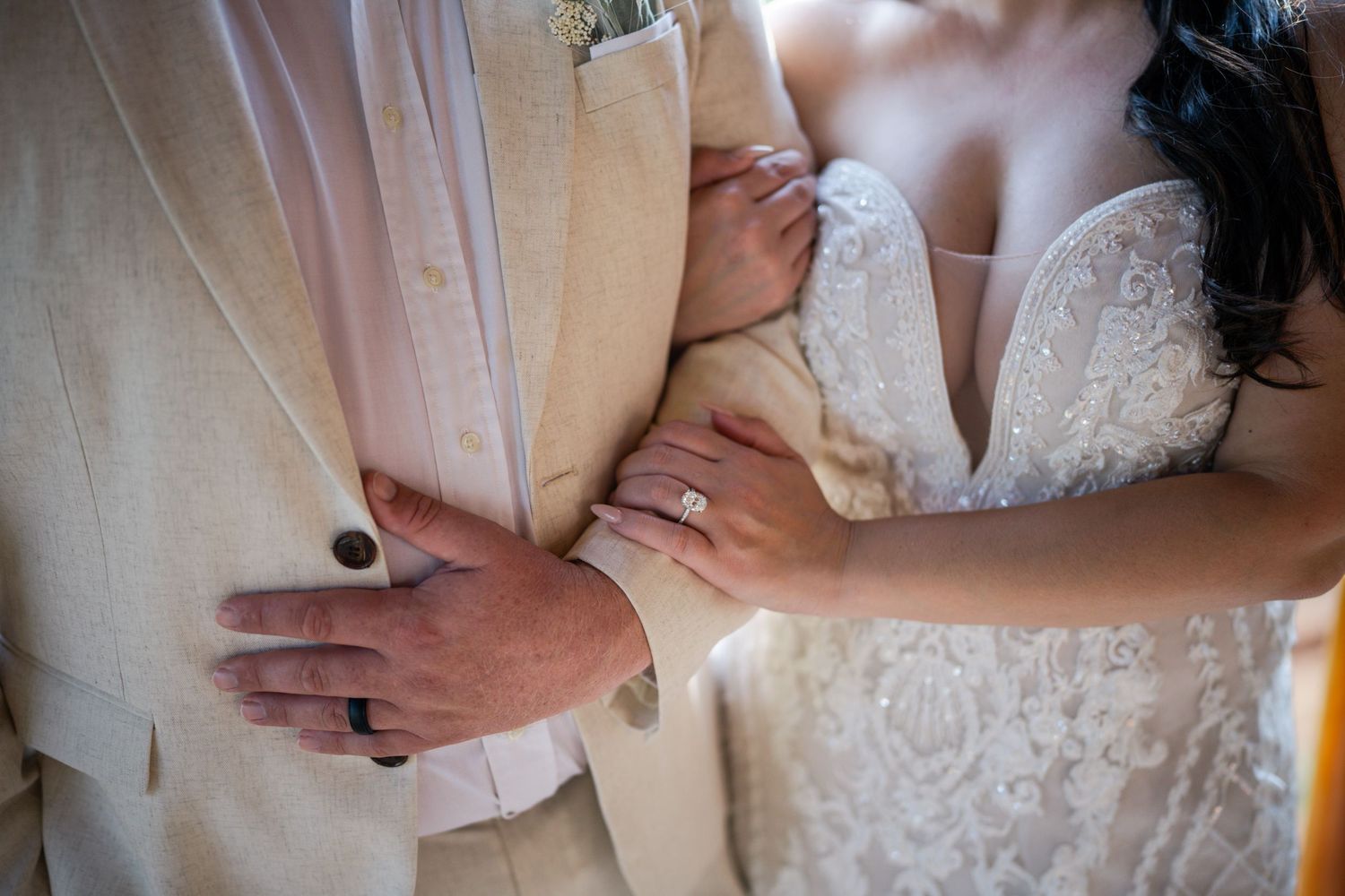 Bride in lace gown embraces groom in tan suit, showcasing wedding rings on their hands.
