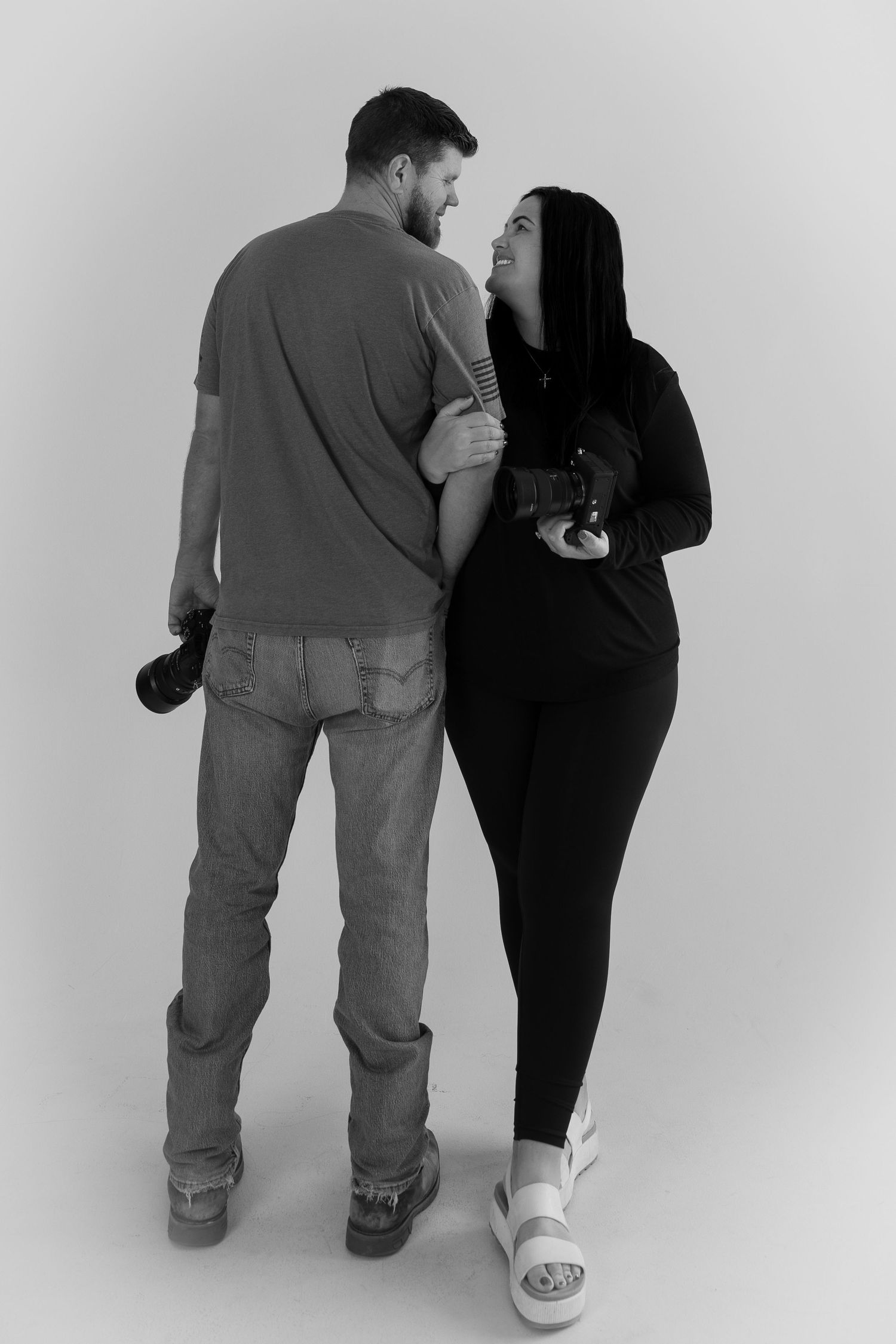 Black and white photo of a couple holding cameras, smiling at each other in a studio setting.