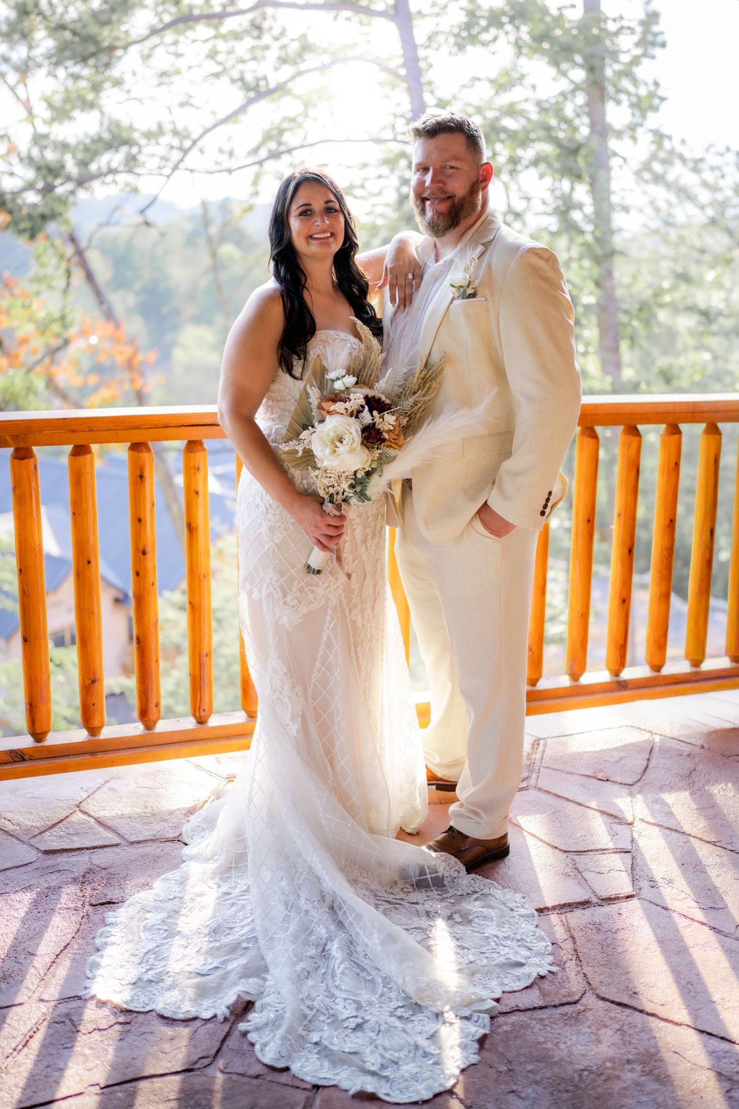 Bride in lace gown and groom in cream suit pose on rustic log cabin deck with mountain views.