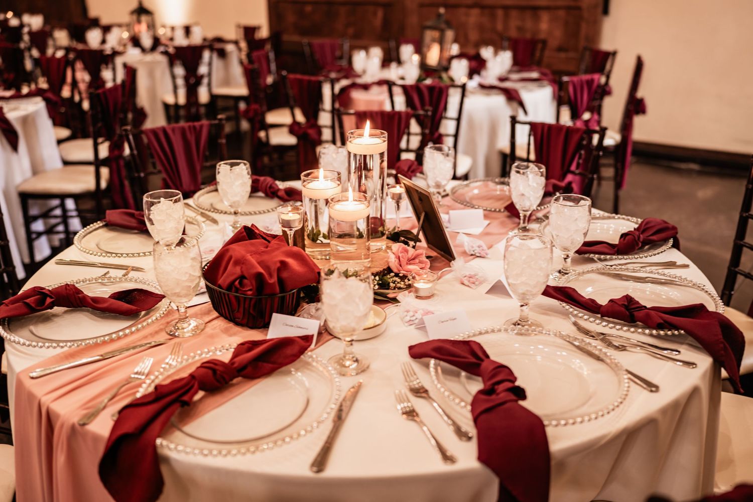 Elegant wedding reception table with burgundy napkins, white linens, gold chargers, candles, and floral centerpiece.