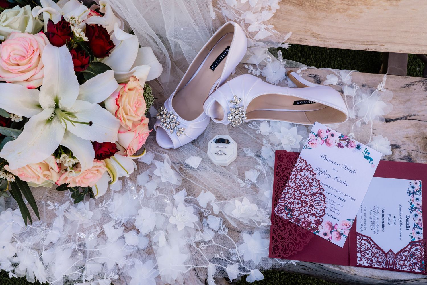 Elegant wedding flat lay featuring white bridal heels, floral bouquet, burgundy laser-cut invitation, and a ring on white petals.