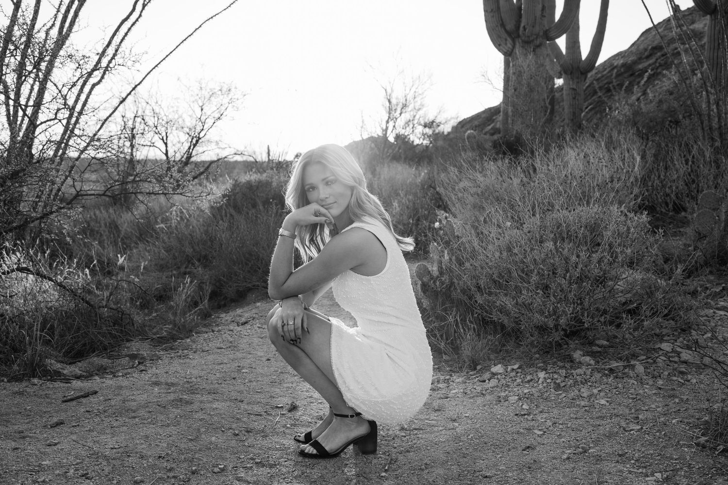 Black and white photo of a blonde woman crouching in a desert landscape with cacti and rocky terrain.
